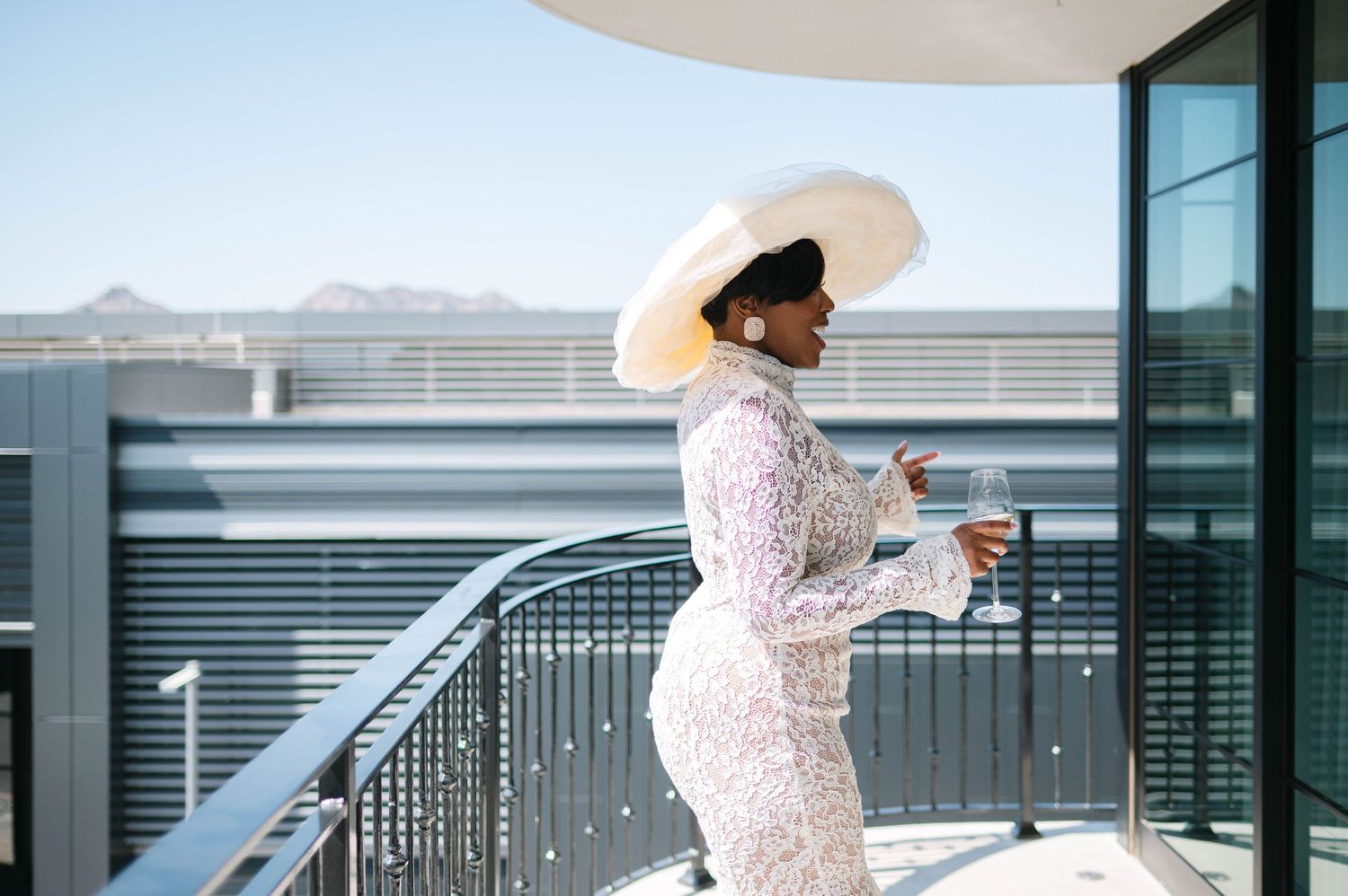 A person in a white lace dress and sun hat poses elegantly on a modern balcony with mountain views in the background.