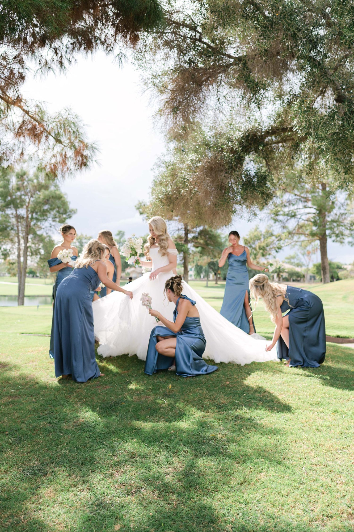 Wedding party members in navy blue attire helping with the bride's dress outdoors.