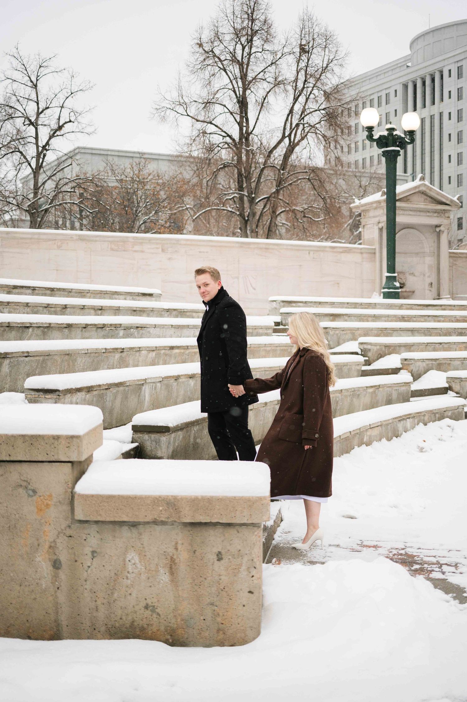 A couple walks along snowy stone steps near a grand building in winter.