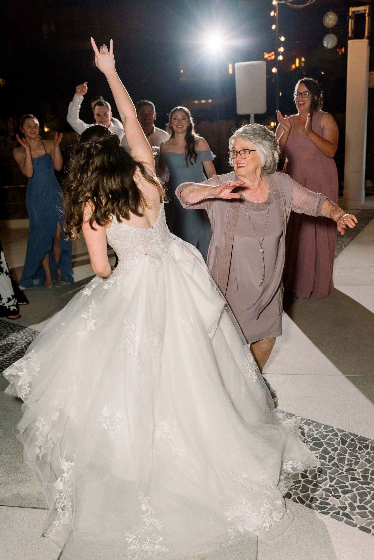 Wedding guests dancing energetically on a patterned floor under string lights at an outdoor reception.