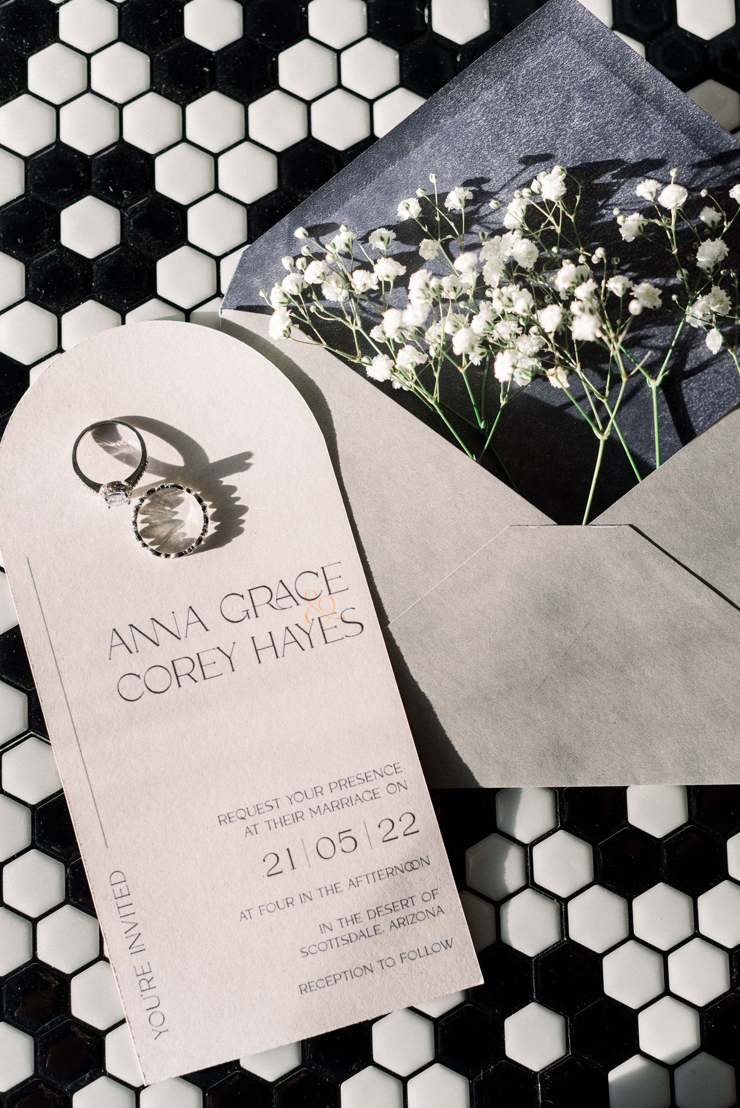 Wedding invitation with elegant black calligraphy on white paper displayed against hexagonal tile with baby's breath flowers.