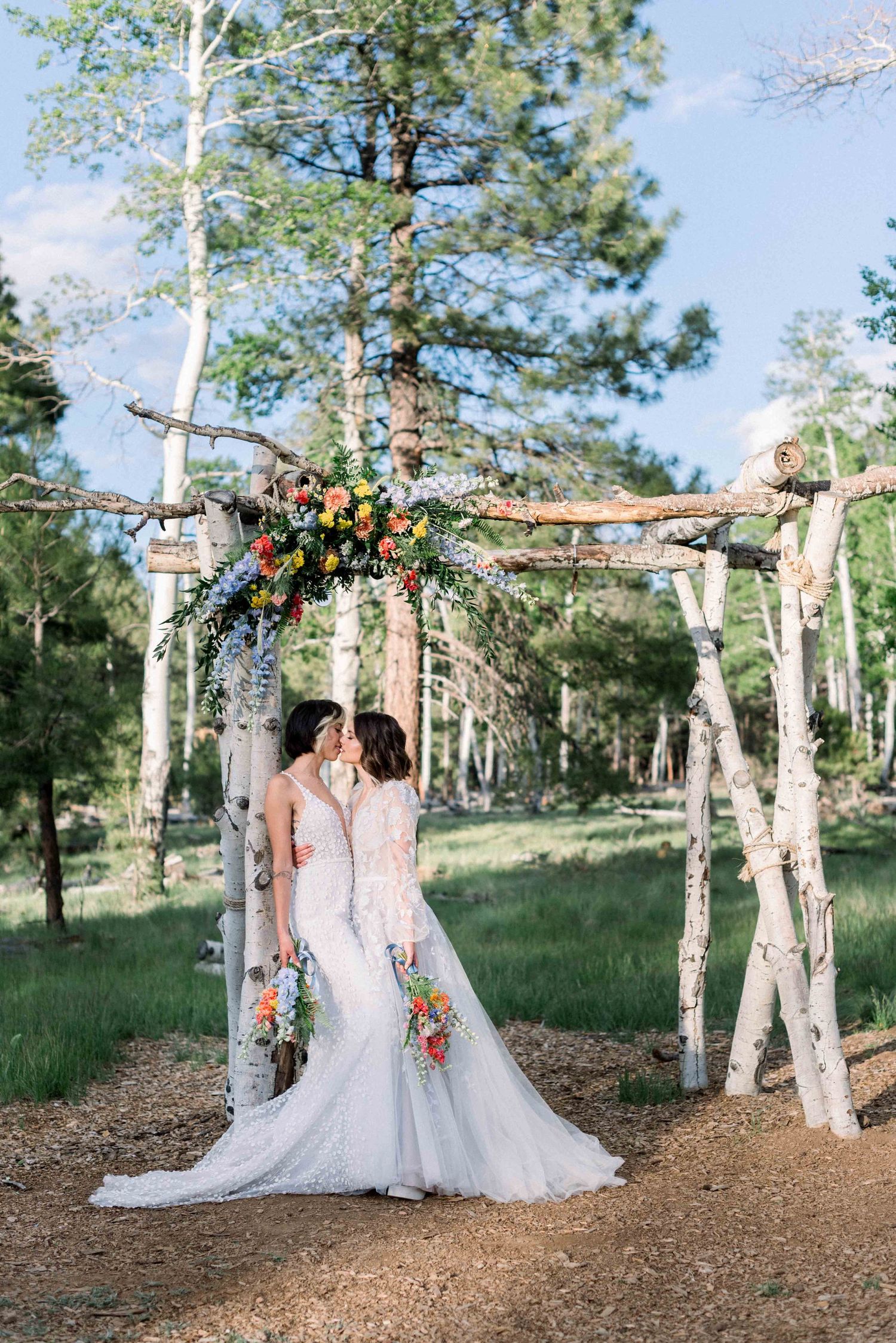 Rustic birch wood wedding arch decorated with colorful flowers in a pine forest.