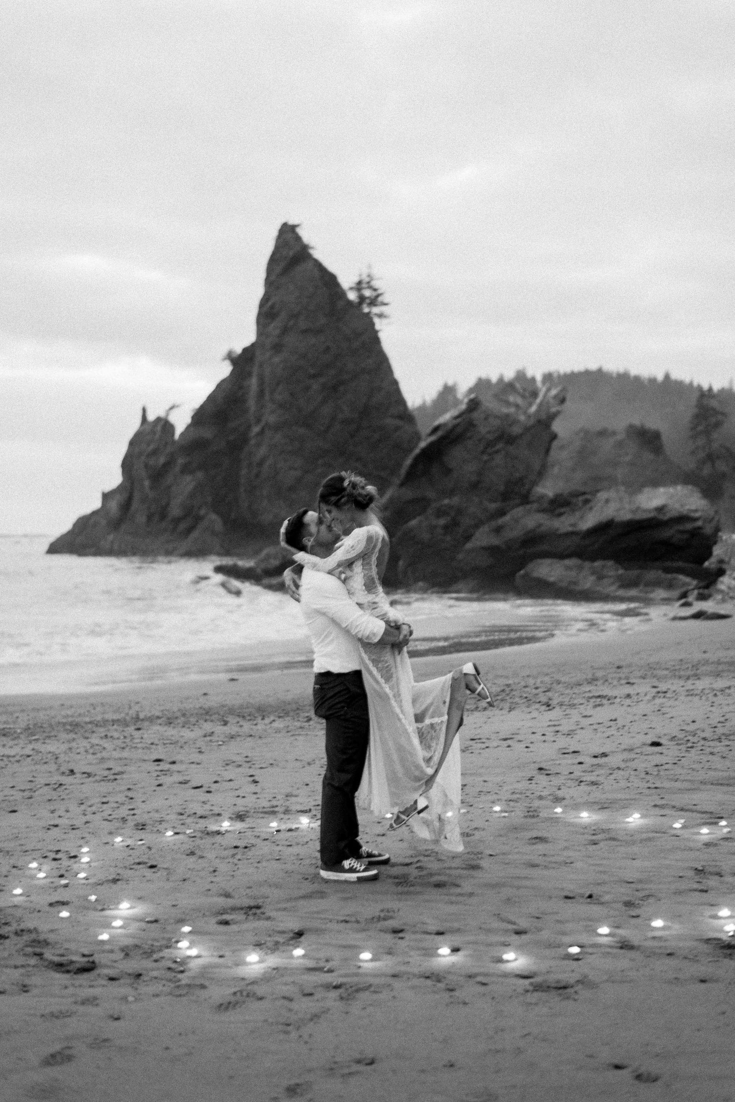 A black and white photo captures a joyful lift during a beach wedding photoshoot.