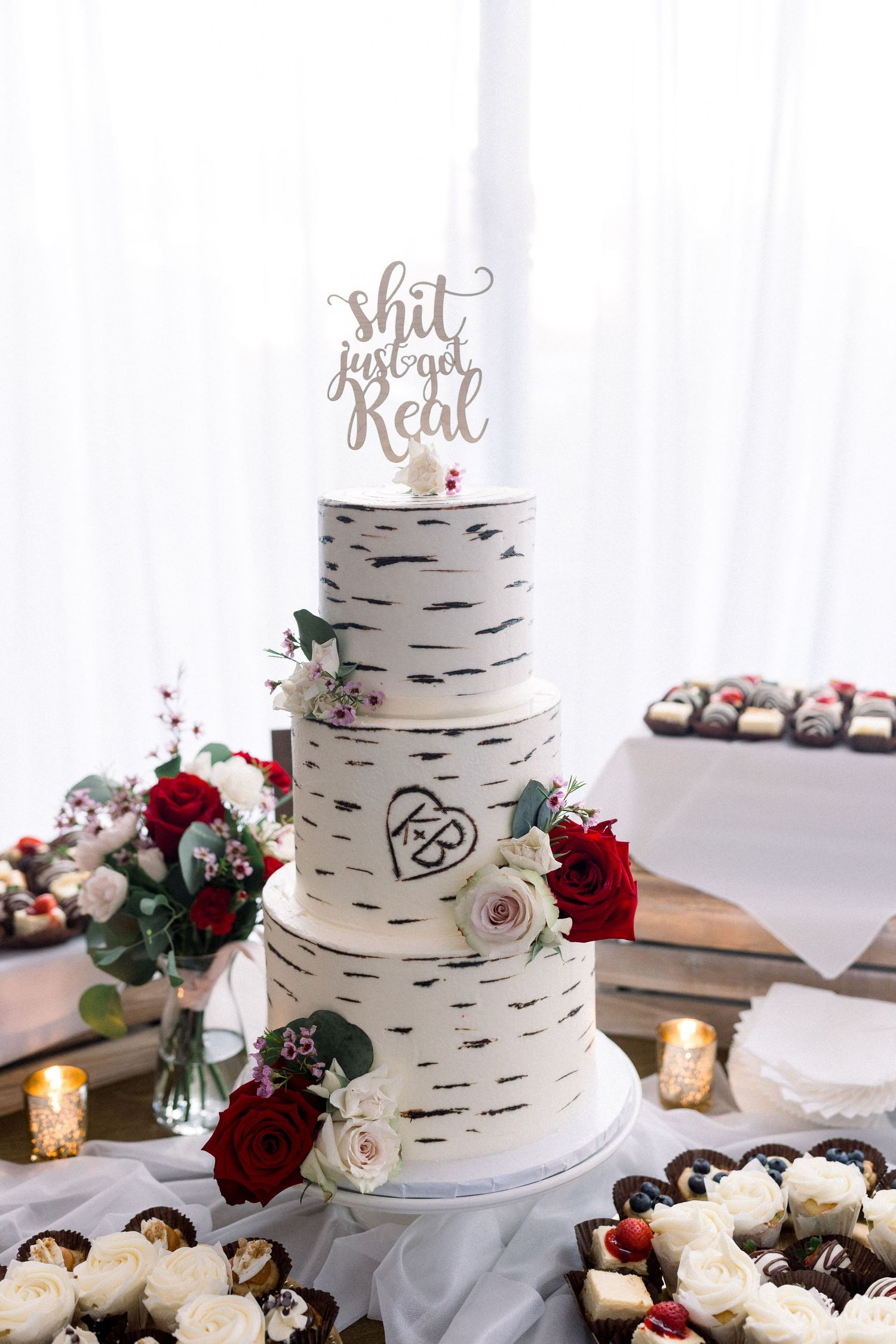 Three-tiered birch bark wedding cake decorated with red roses and a gold cake topper.