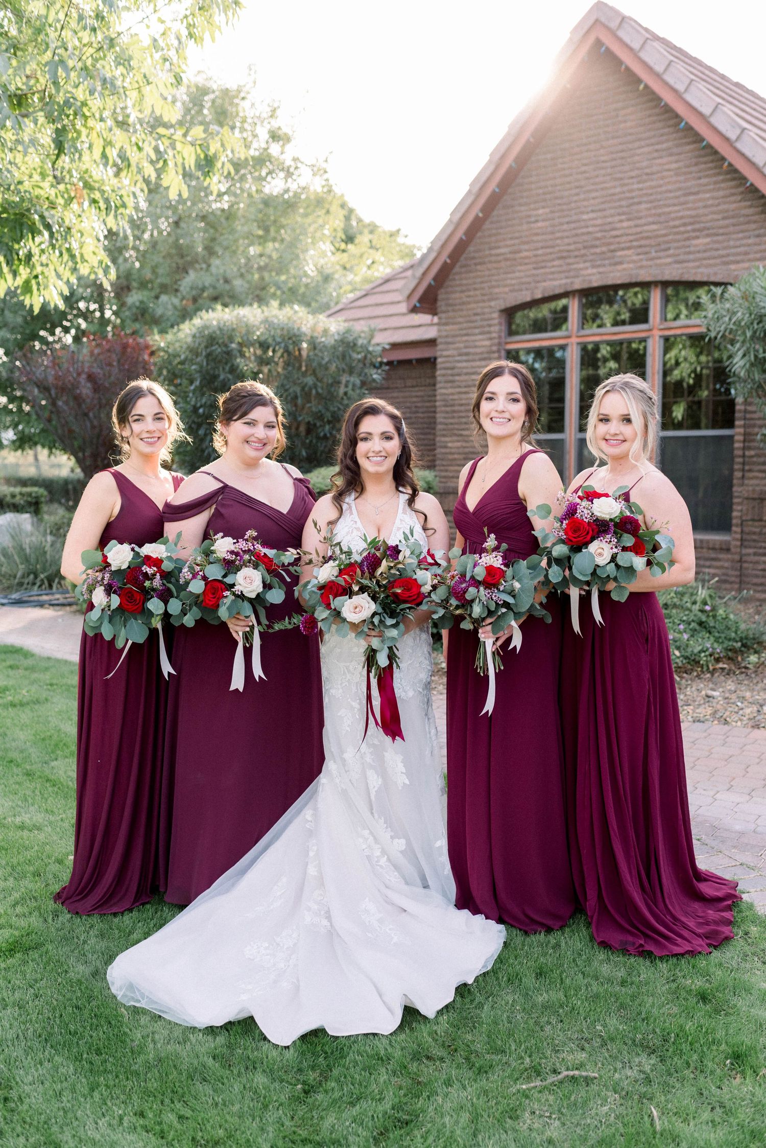Bridal party in burgundy dresses poses with bouquets featuring red and white flowers.