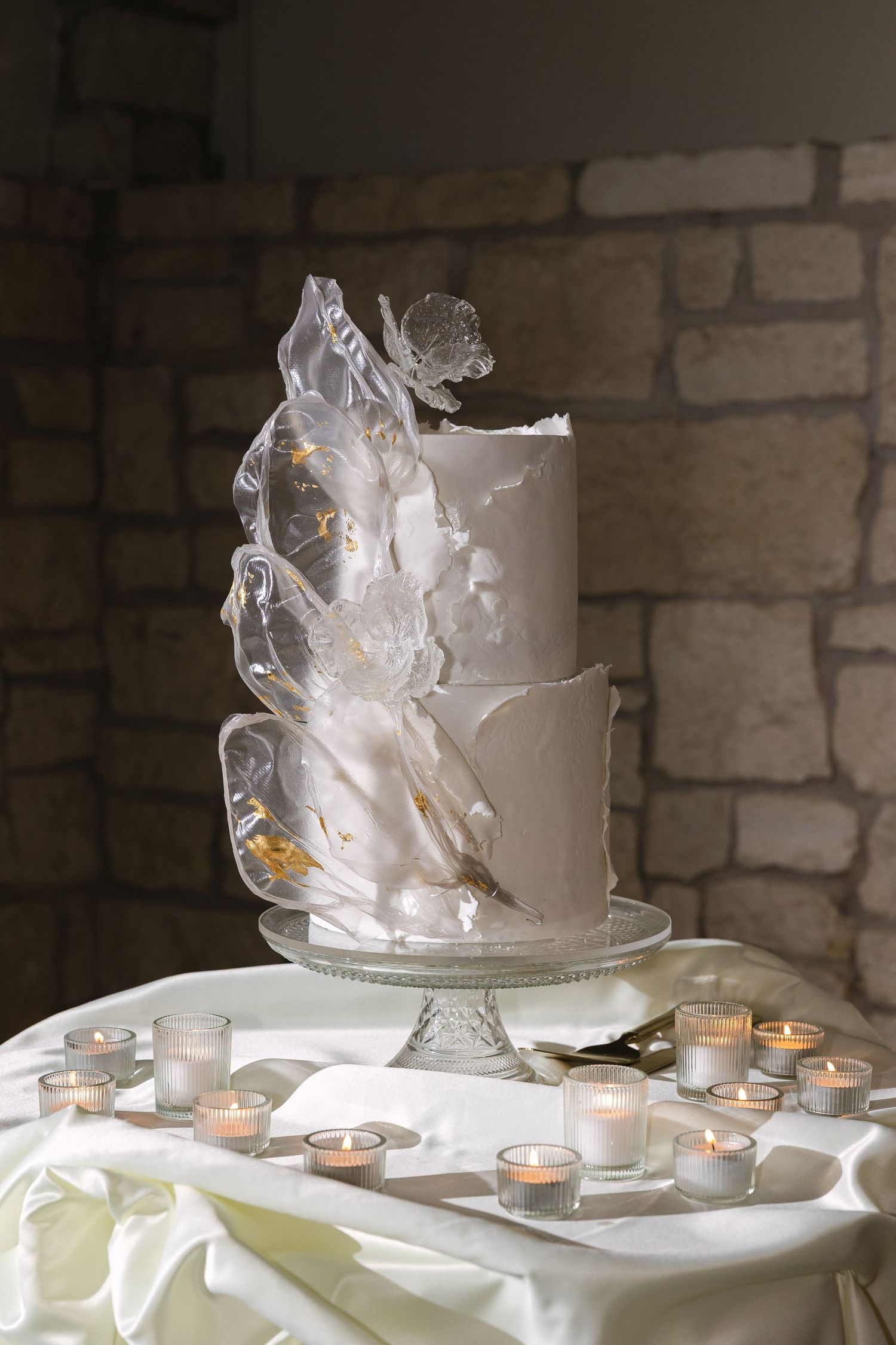 An elegant white wedding cake display with glowing tea lights on a stone-walled backdrop.