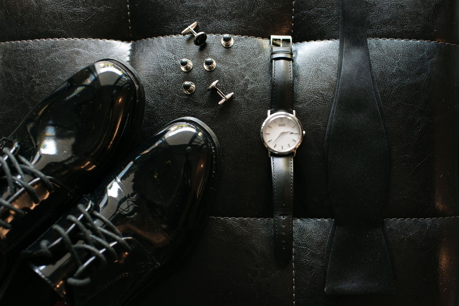 A classic wristwatch with leather strap placed next to polished black leather dress shoes on dark surface.