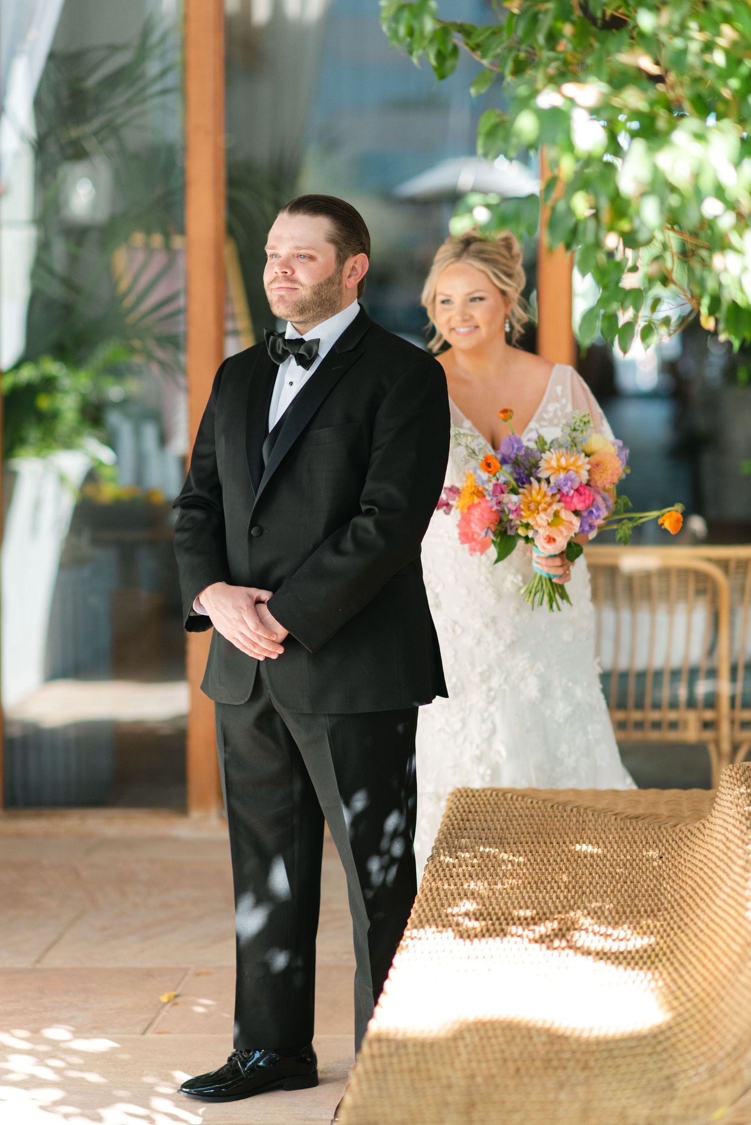 A bride and groom share a joyful moment on a sunny patio with woven furniture and dappled light through greenery.