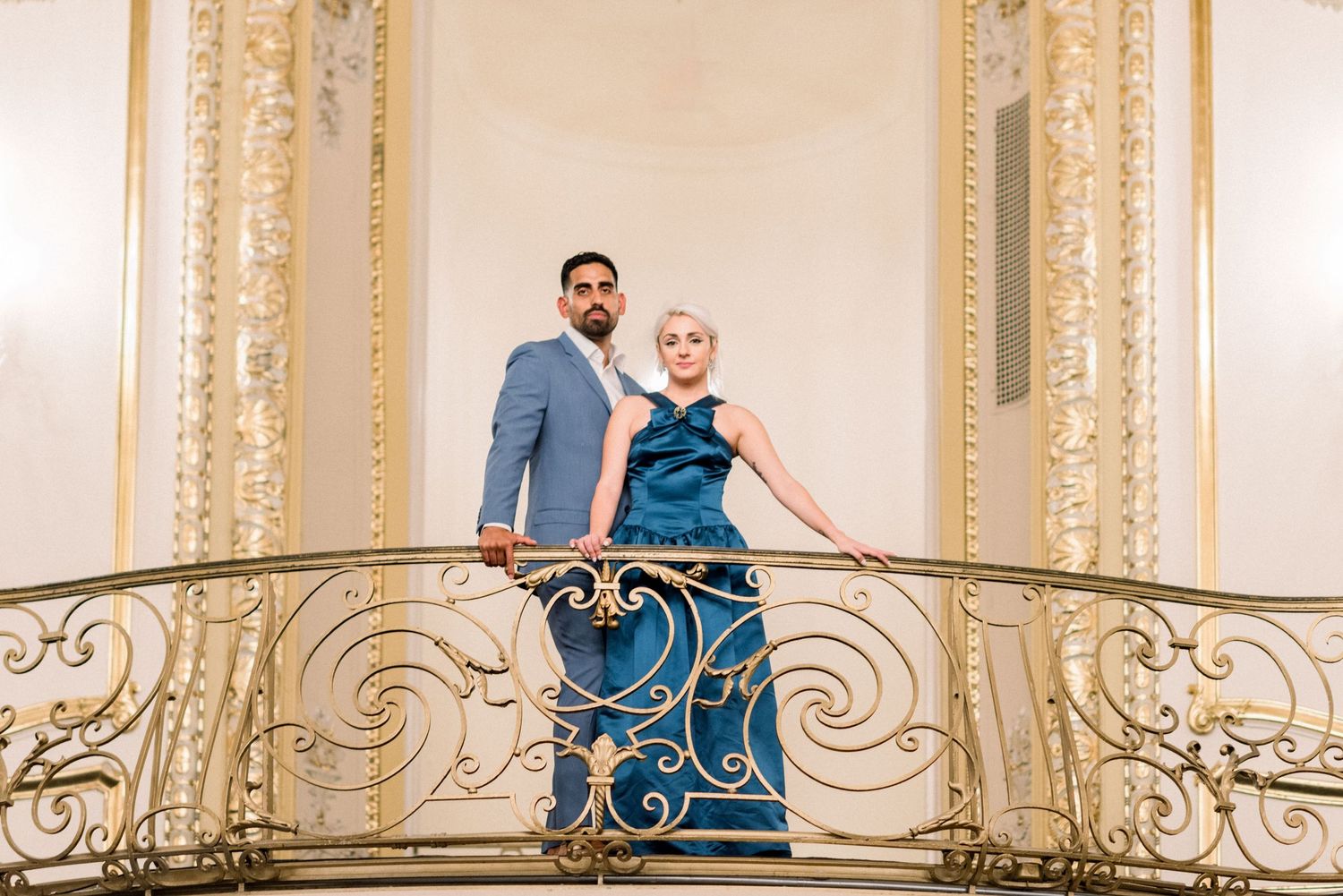 Elegant pair stands on decorative gold balcony in historic venue.