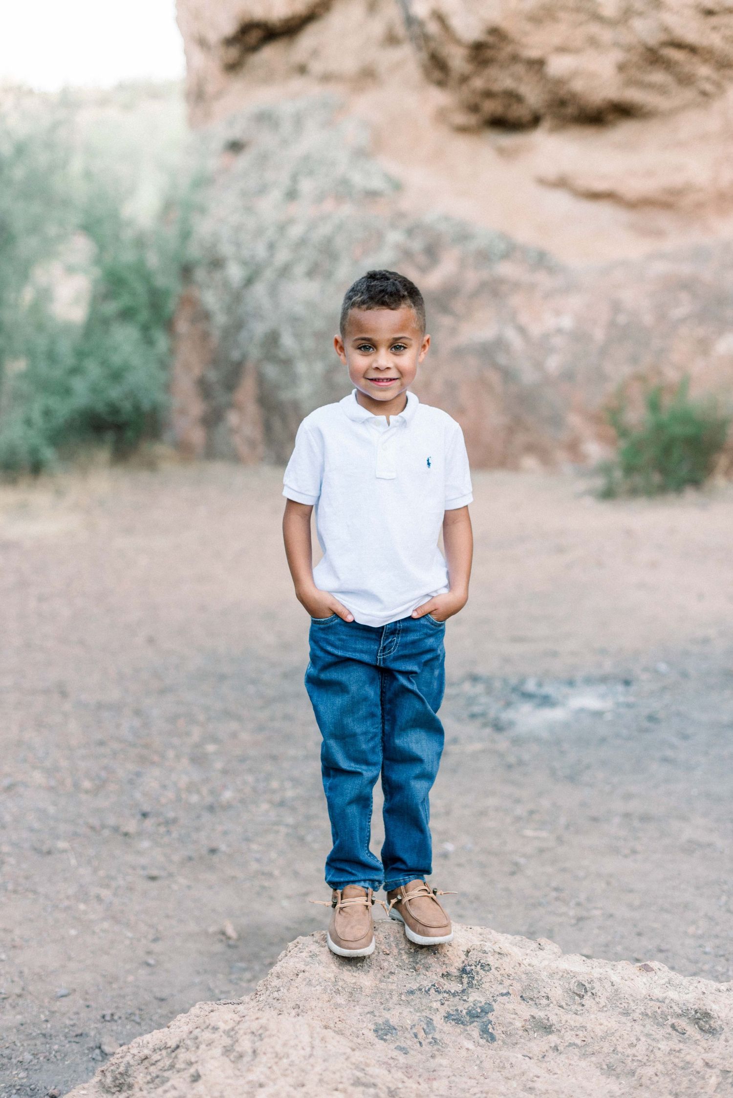 A young child in white polo and jeans stands confidently on a rock formation in the desert.