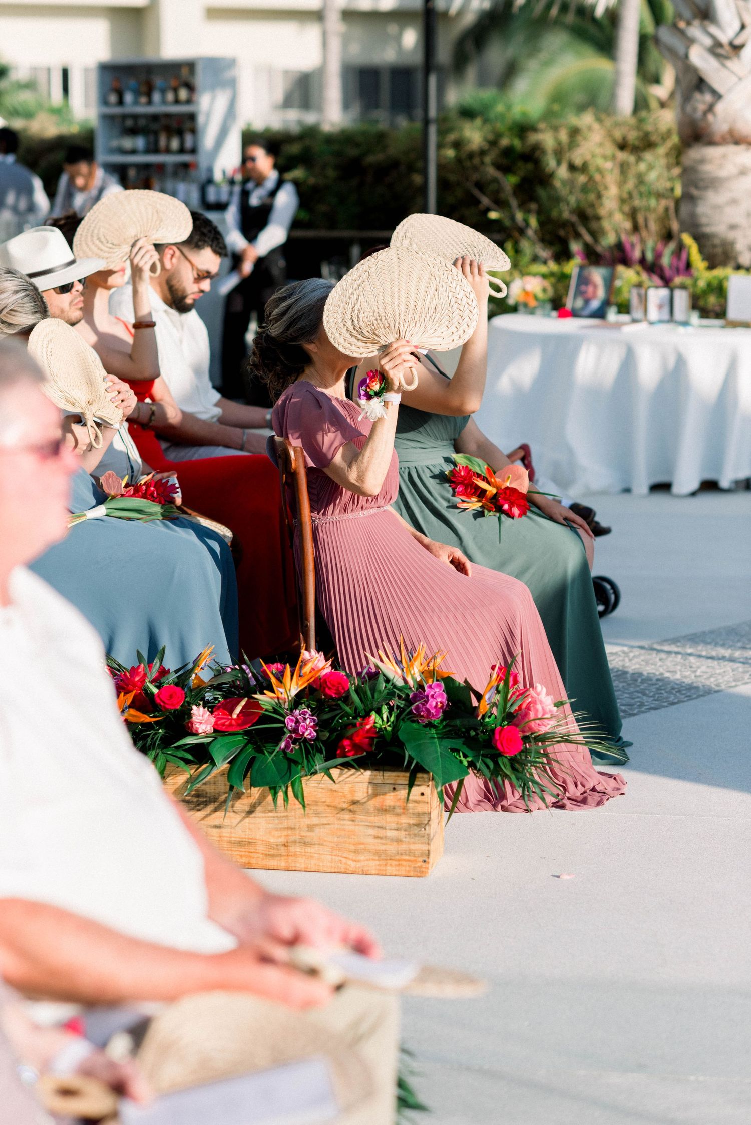 Wedding guests in sun hats seated outdoors with floral decorations and a tropical backdrop.