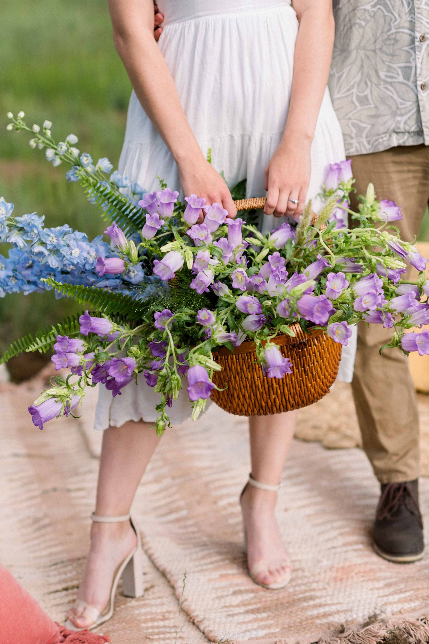 Close-up of wicker basket filled with purple and blue wildflowers held against white dress.