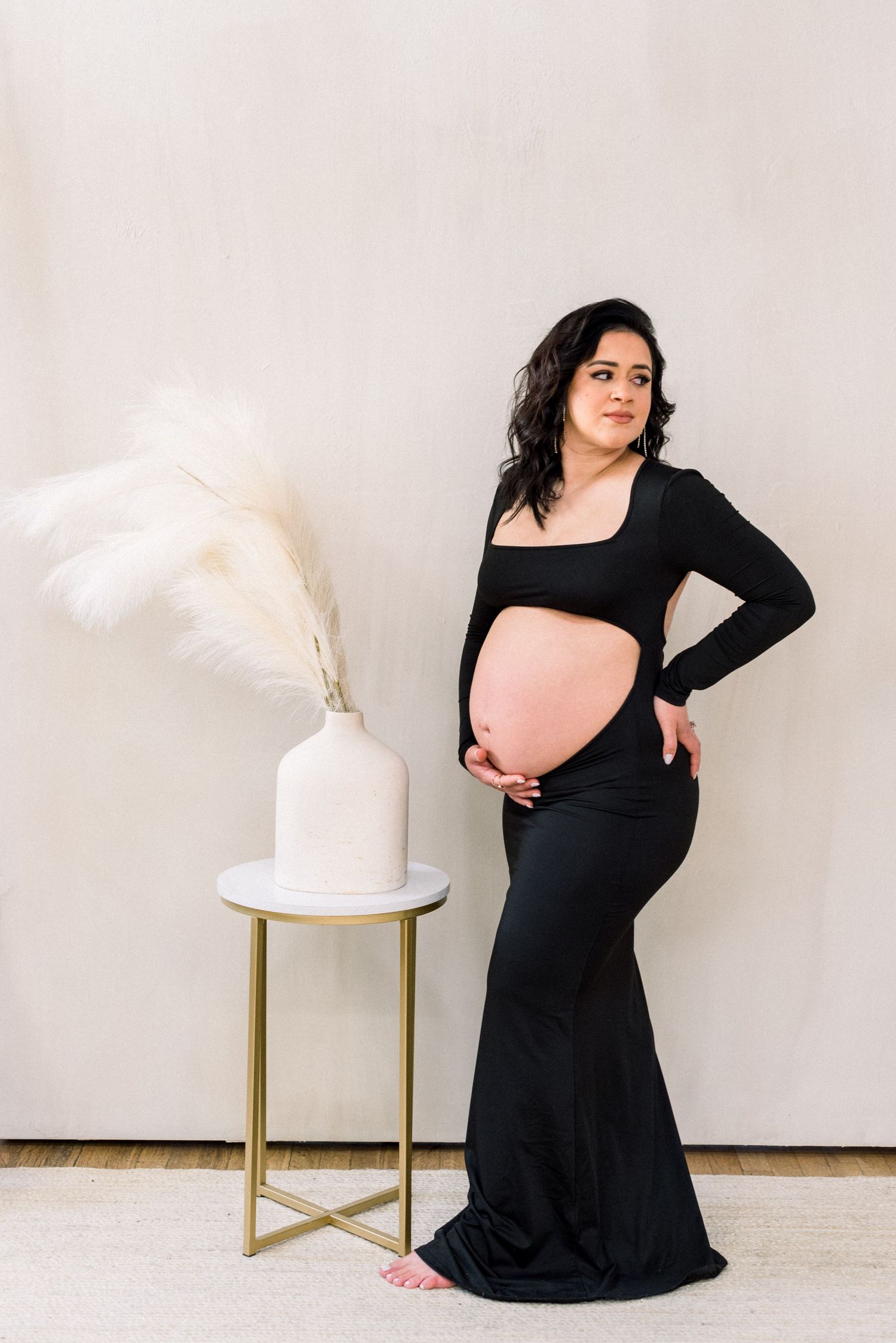 Maternity photo of a pregnant woman in a black dress posing beside a white vase with pampas grass.