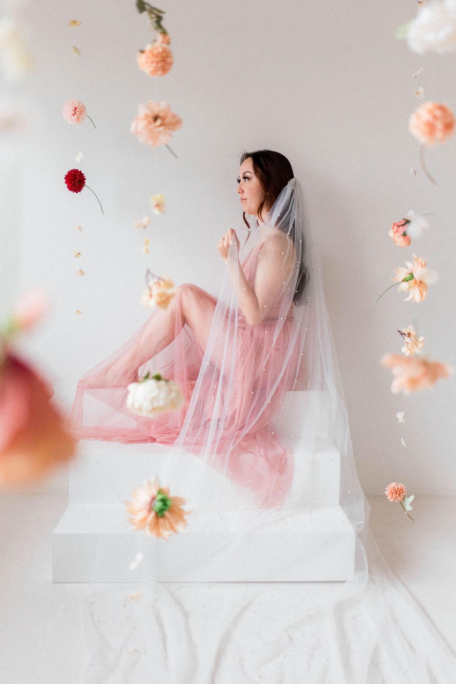 Dreamy portrait with floating flowers and veil surrounding figure in pink dress on white platform.