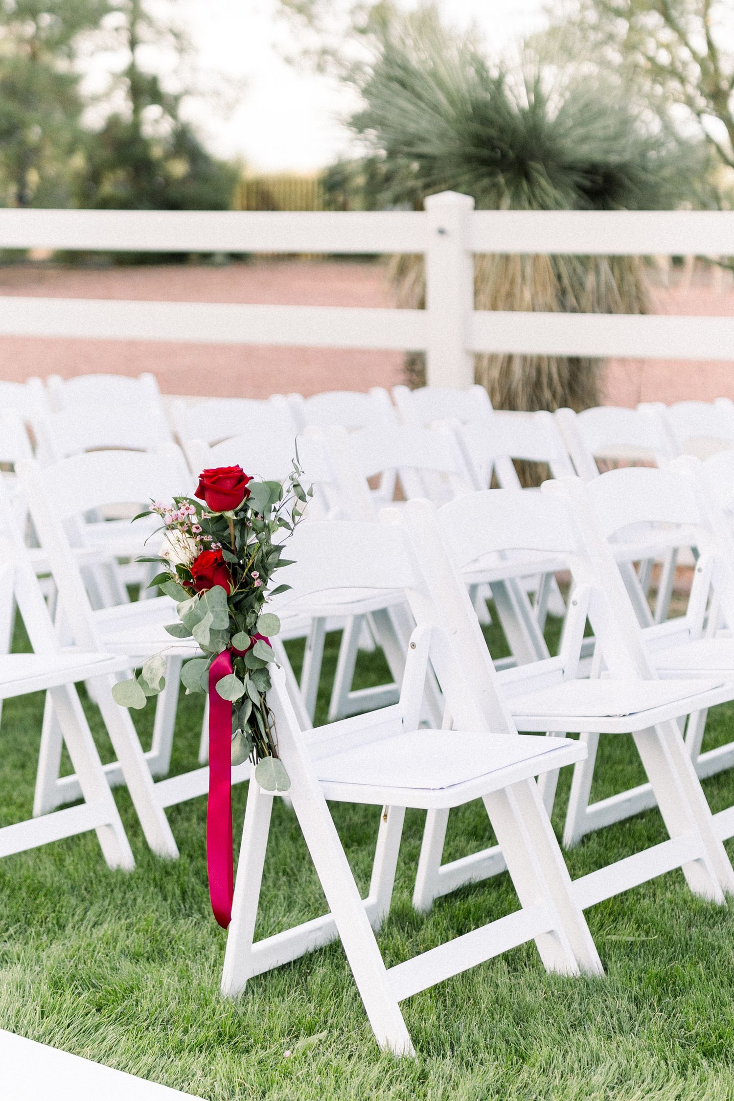 White wedding chairs adorned with red roses and burgundy ribbons in an outdoor garden setting.