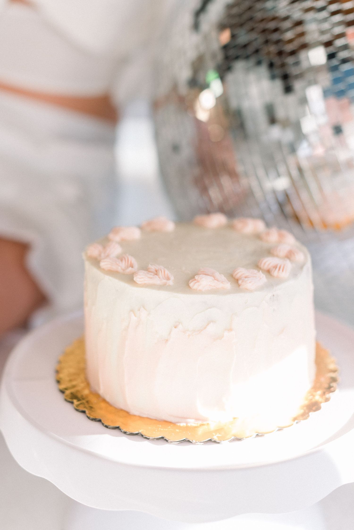 White cake with delicate pink frosting decorations on a gold cake board with disco ball in background.