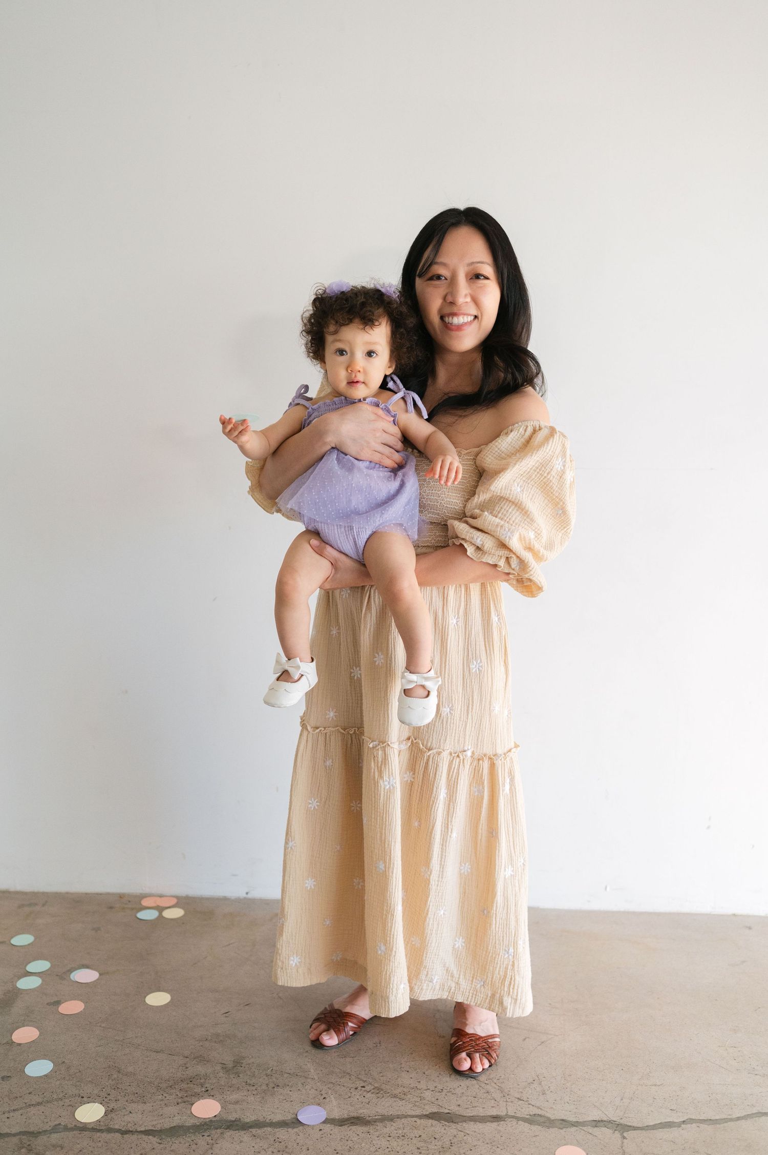 A parent and child in matching beige dresses stand together against a white wall with pastel confetti on the floor.