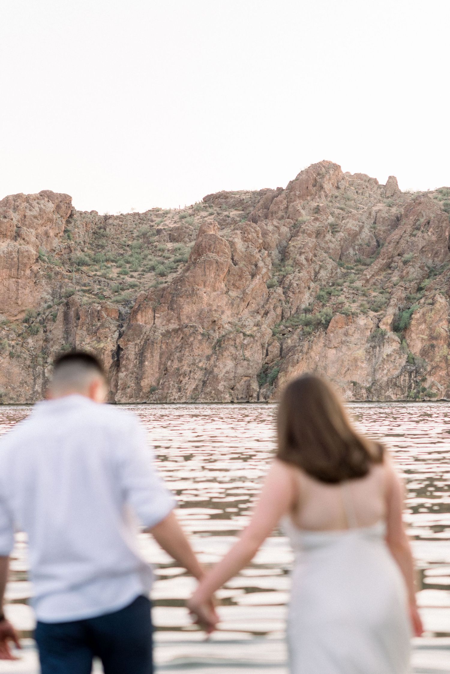 A couple holds hands while overlooking a scenic canyon lake with towering red rock cliffs in the background.