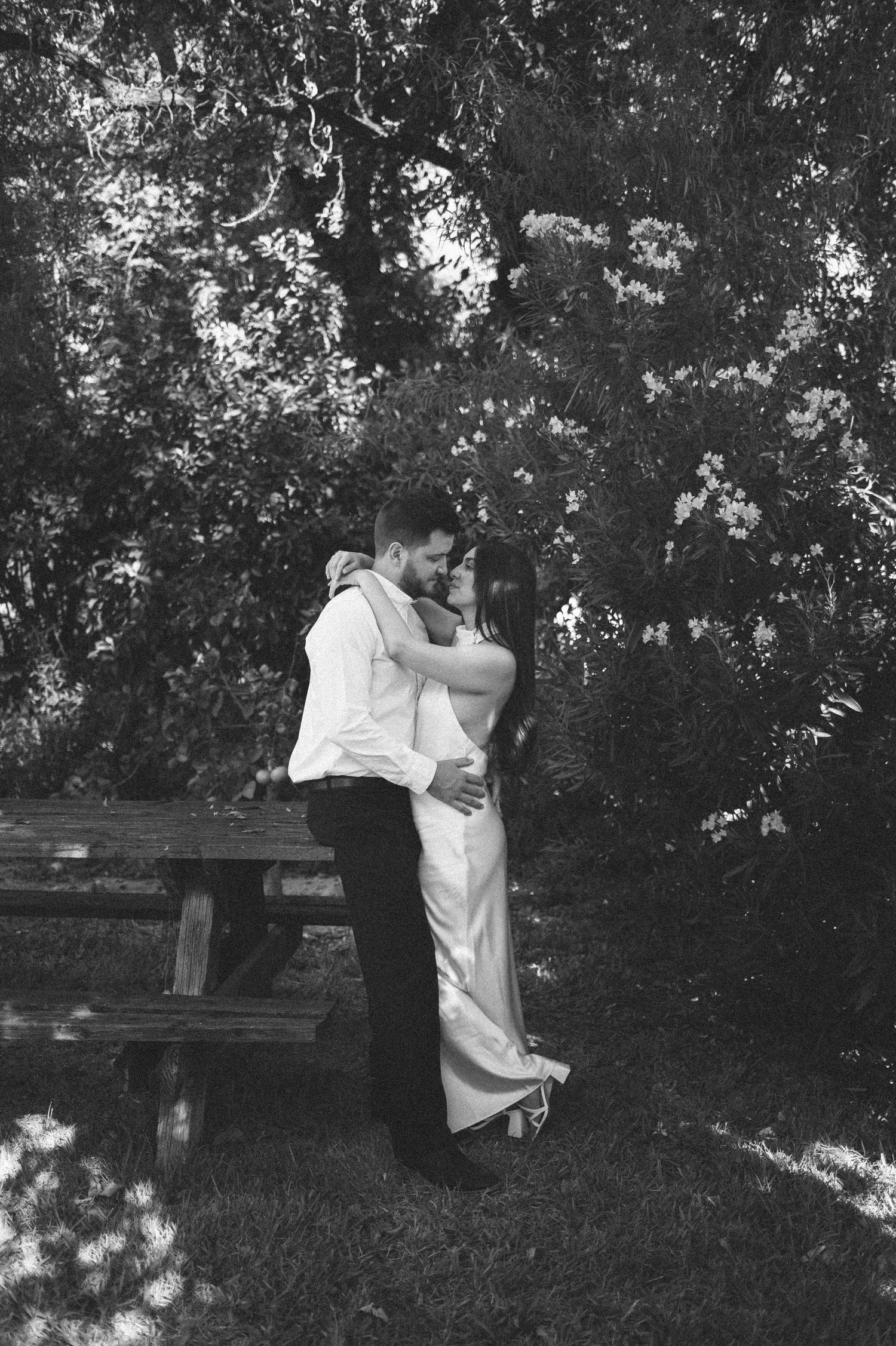 A couple embraces in a romantic moment on a grassy lawn with trees in the background, captured in black and white.