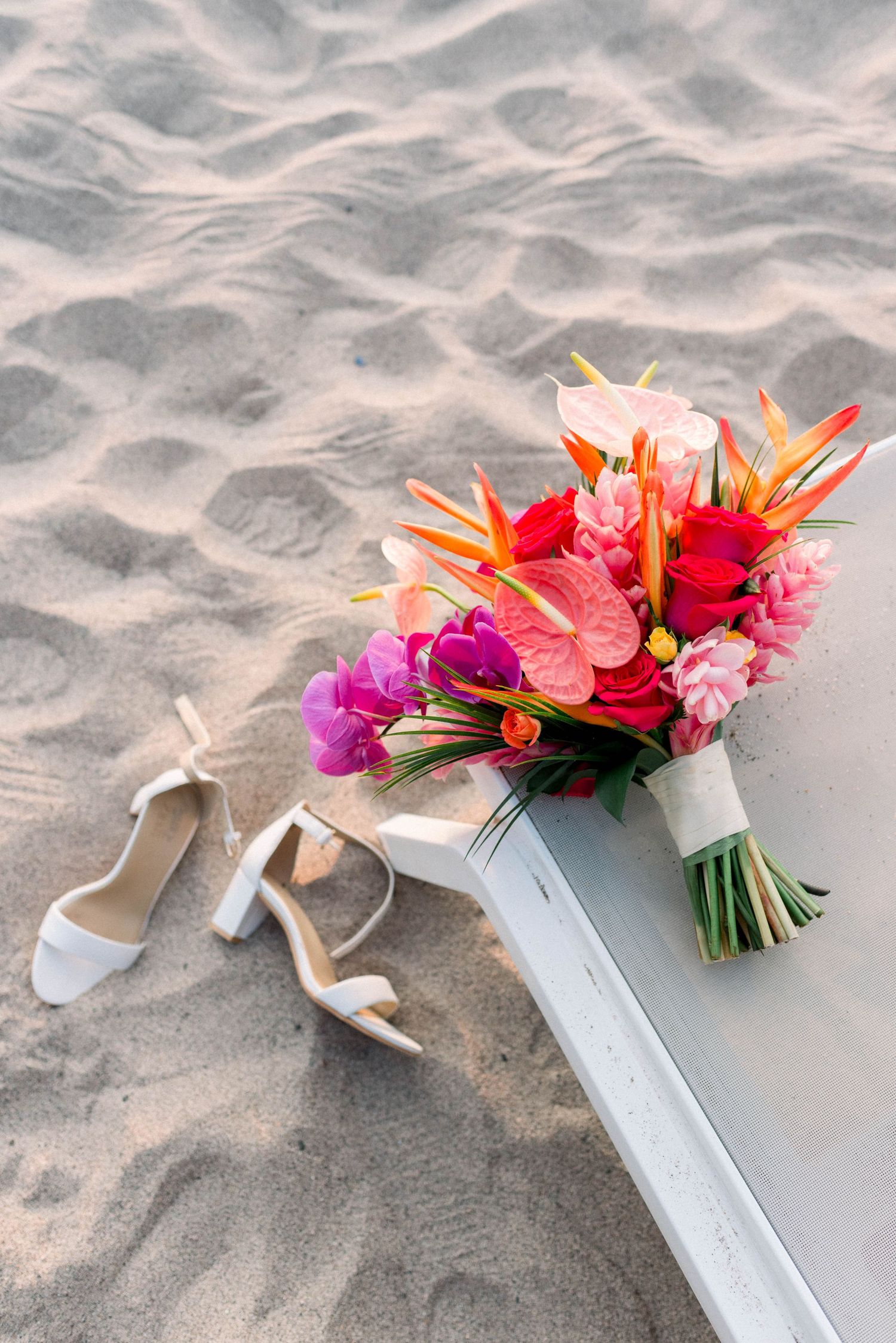 Bridal bouquet with tropical flowers and wedding shoes arranged artistically on sandy beach.