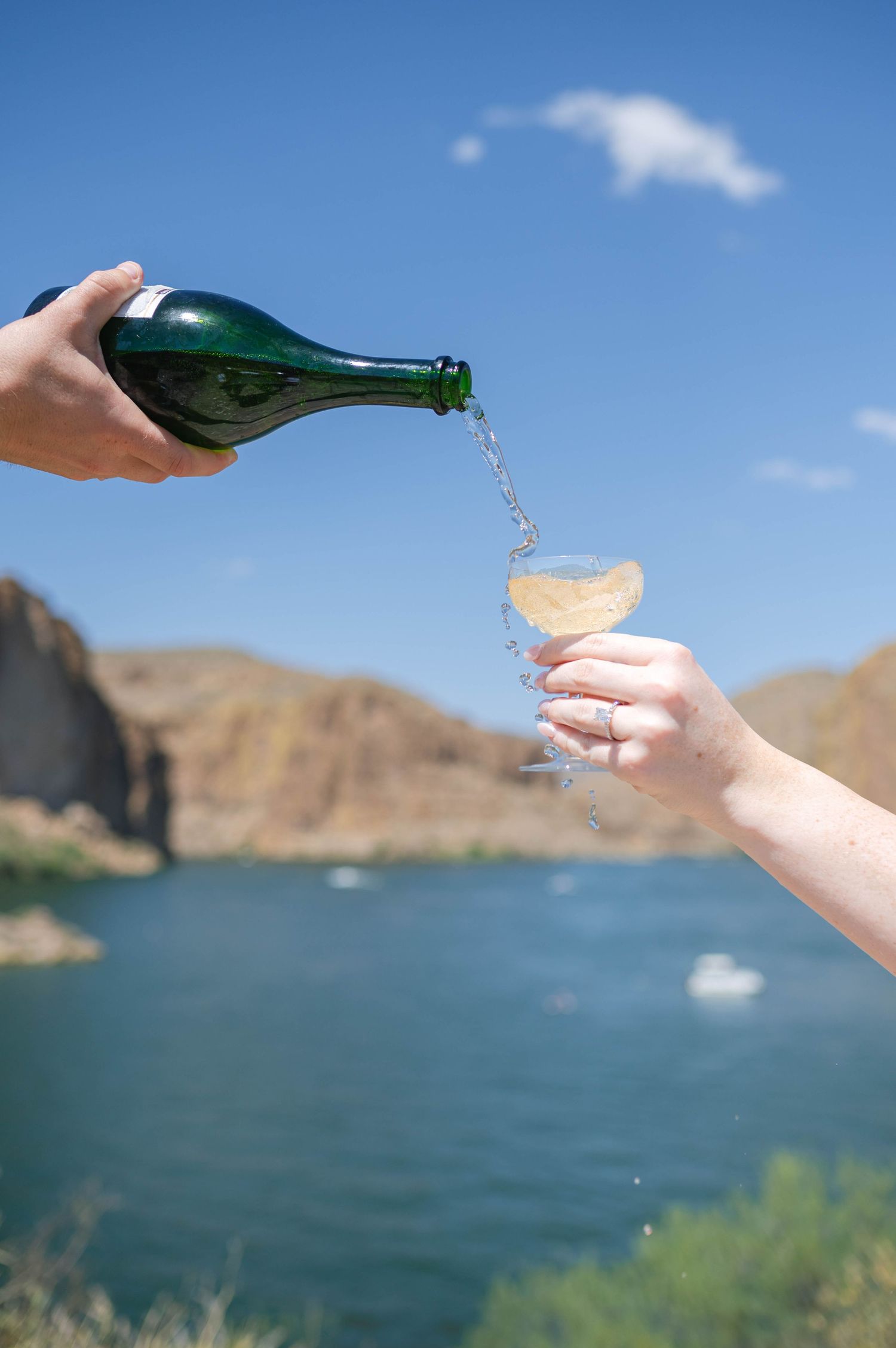 Two hands pouring champagne into glasses against a scenic lake and mountain backdrop.