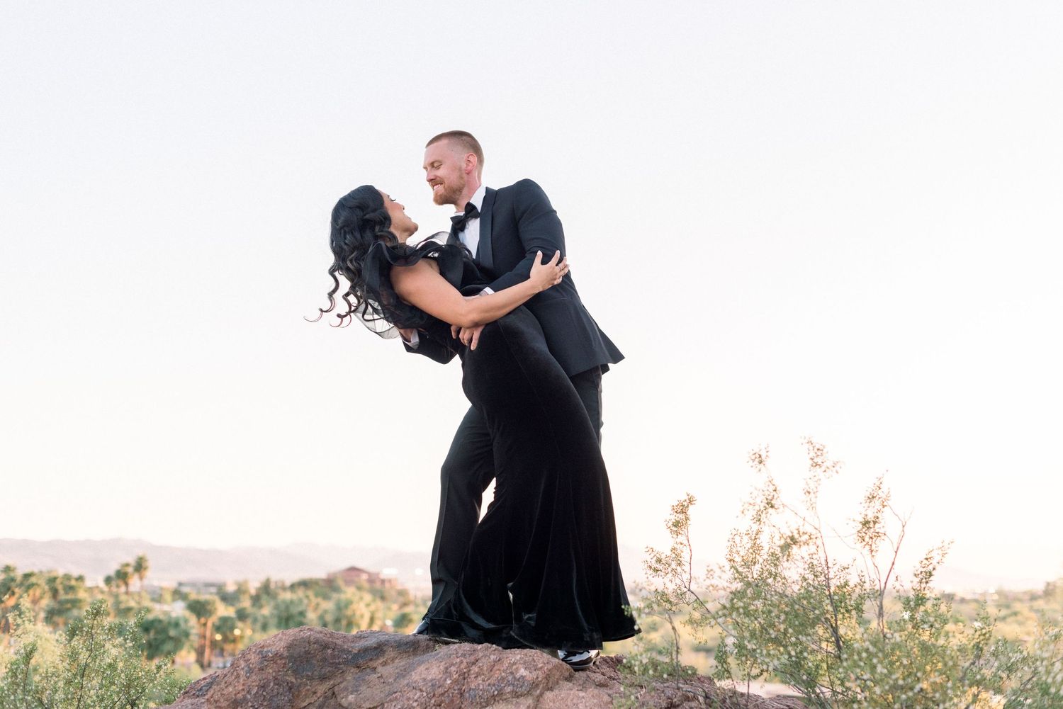 A romantic couple in formal black attire embraces against a desert landscape during sunset.