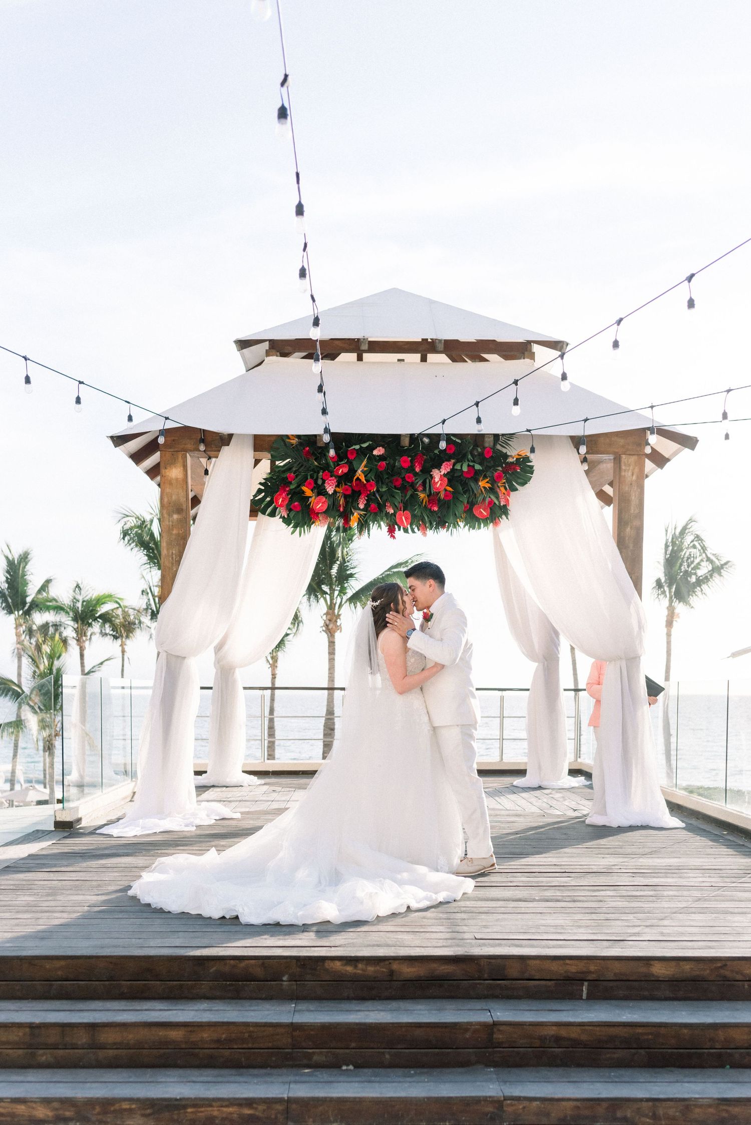 A romantic wedding ceremony under a white draped gazebo decorated with red flowers and string lights by the ocean.