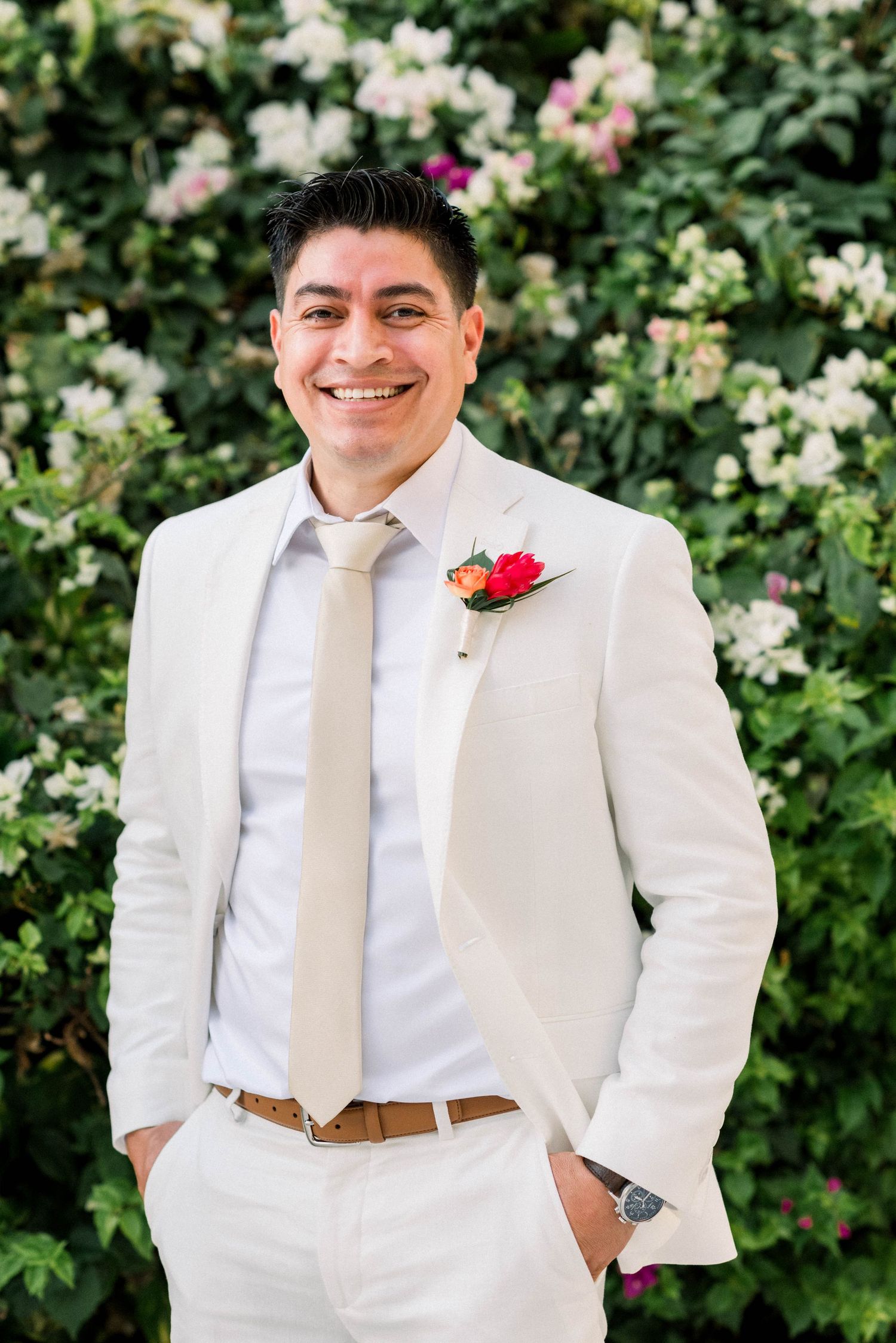 Groom in light colored suit with red boutonniere smiles against floral garden backdrop.