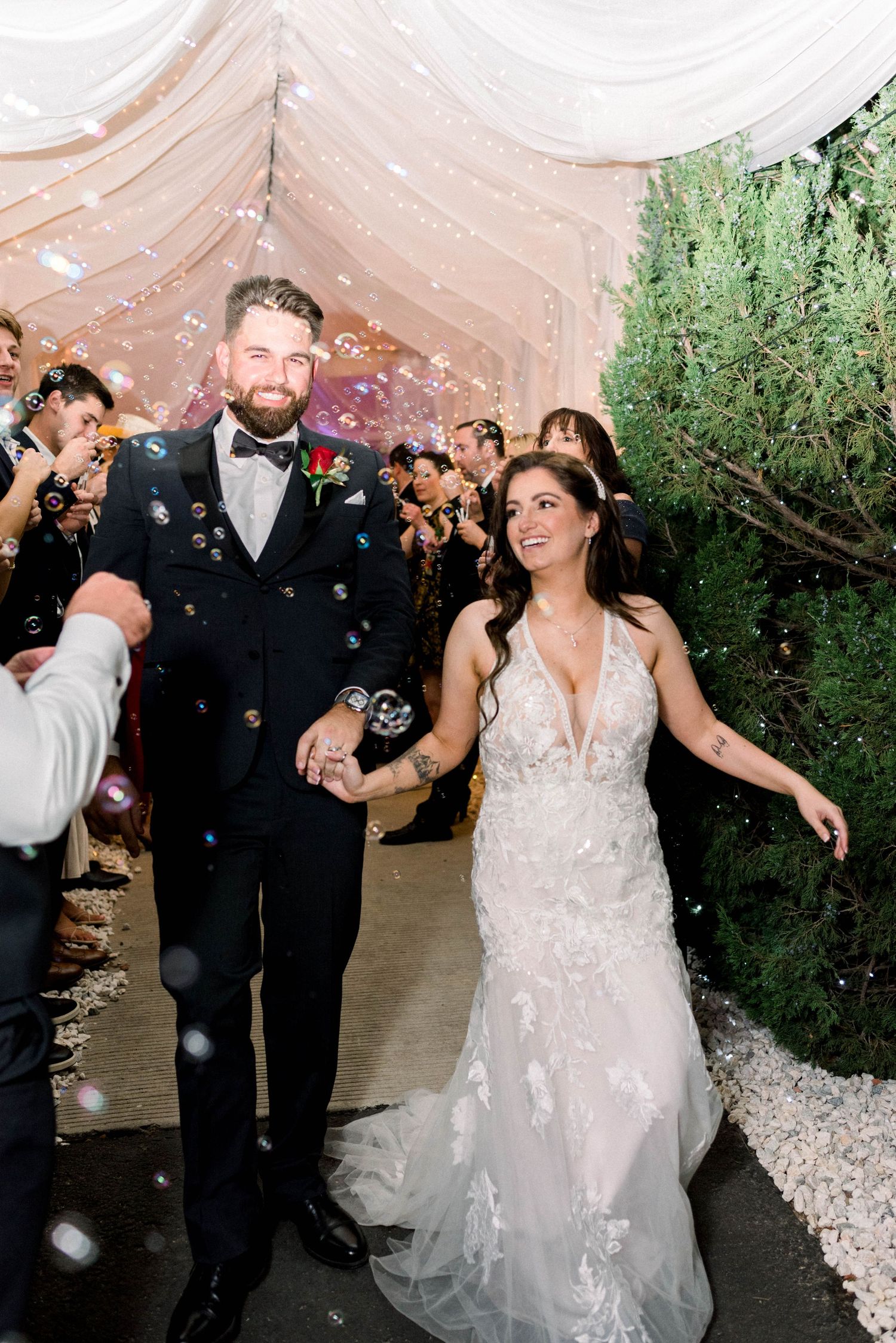Newlyweds walk through confetti shower during wedding reception exit under white tent with greenery.