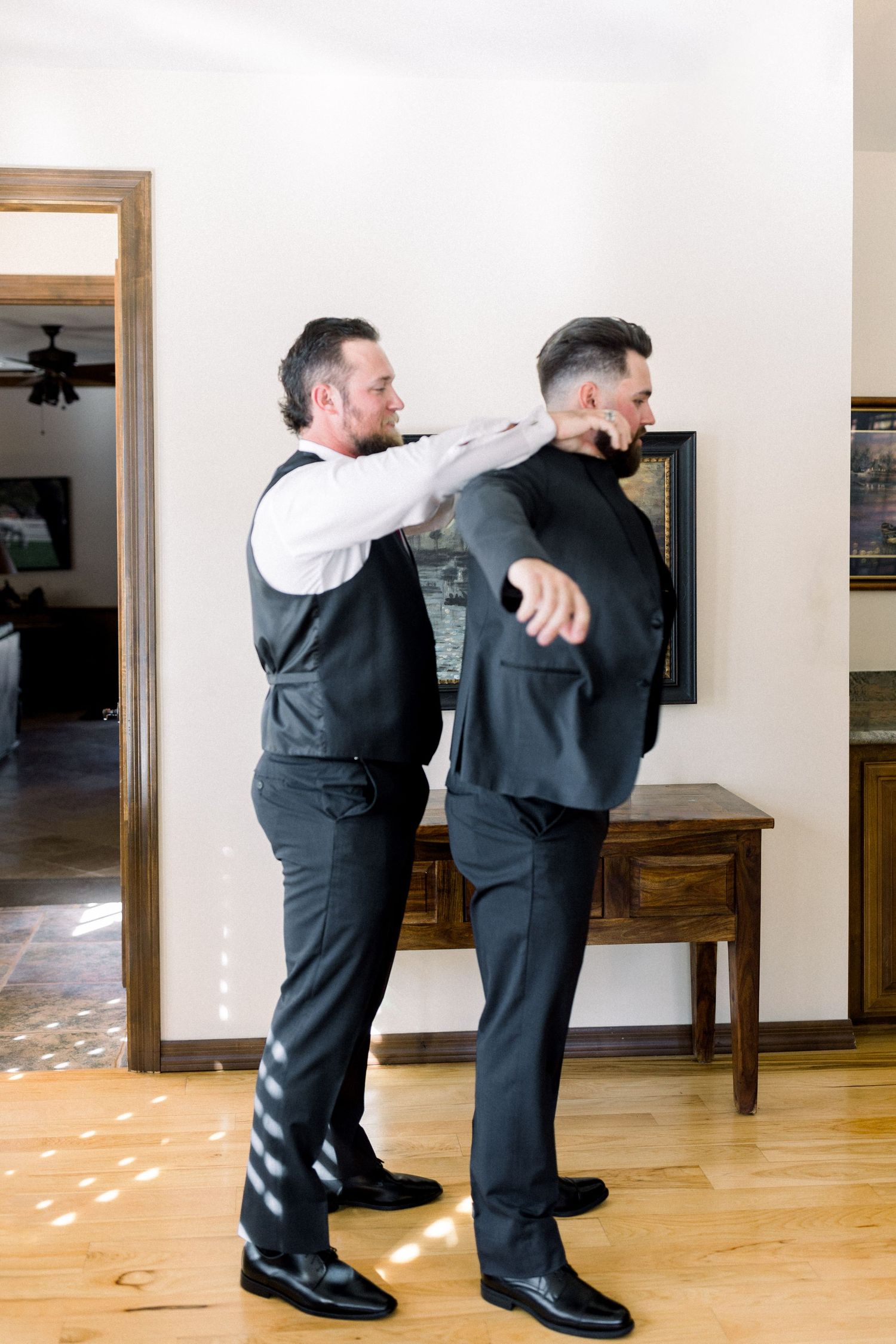 Best man helps groom get ready by adjusting his suit jacket in a sunlit room with hardwood floors.