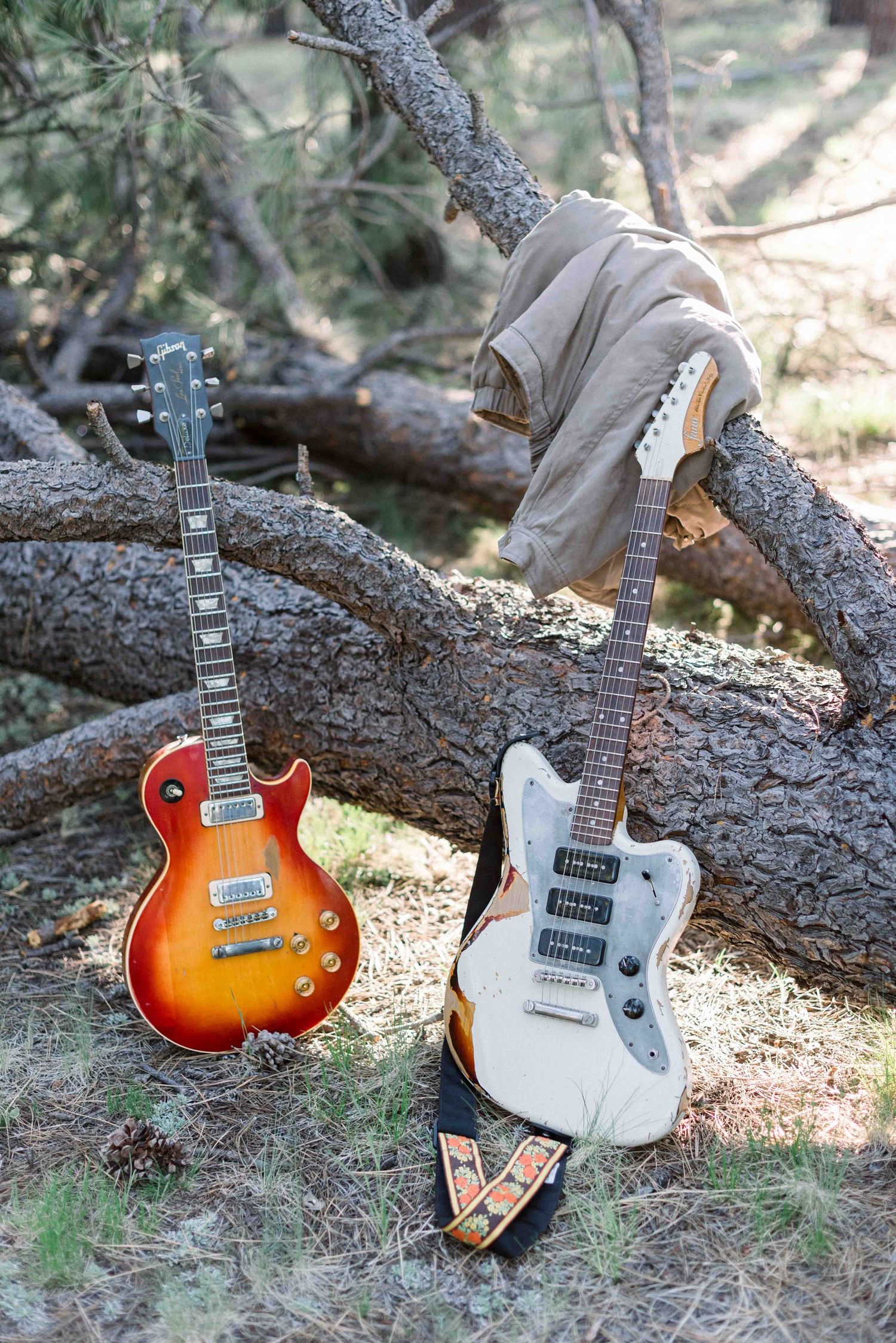 Two electric guitars, one sunburst and one white, displayed on a fallen tree branch in nature.