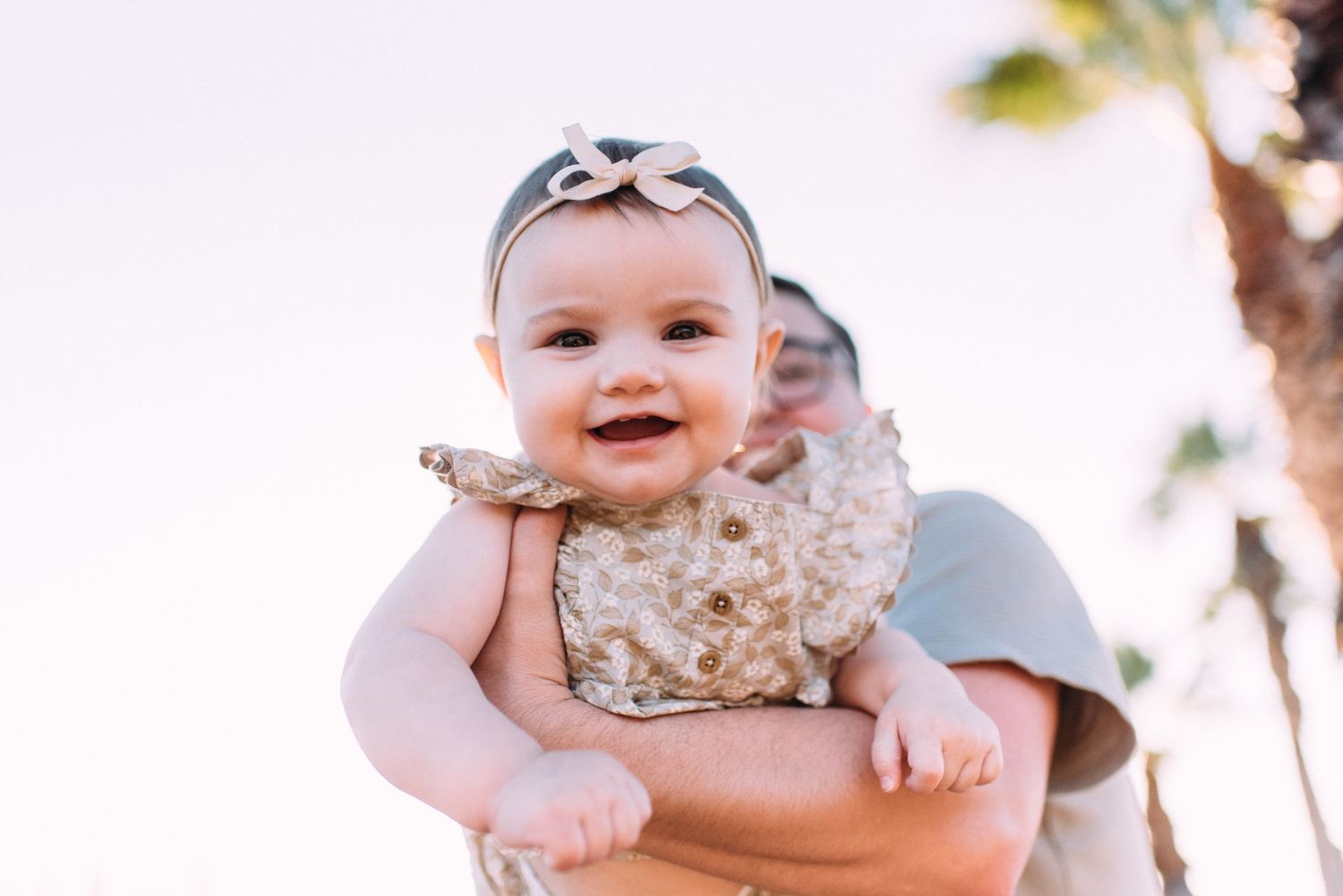 A smiling baby in a floral outfit and bow headband is held up against a bright outdoor background.