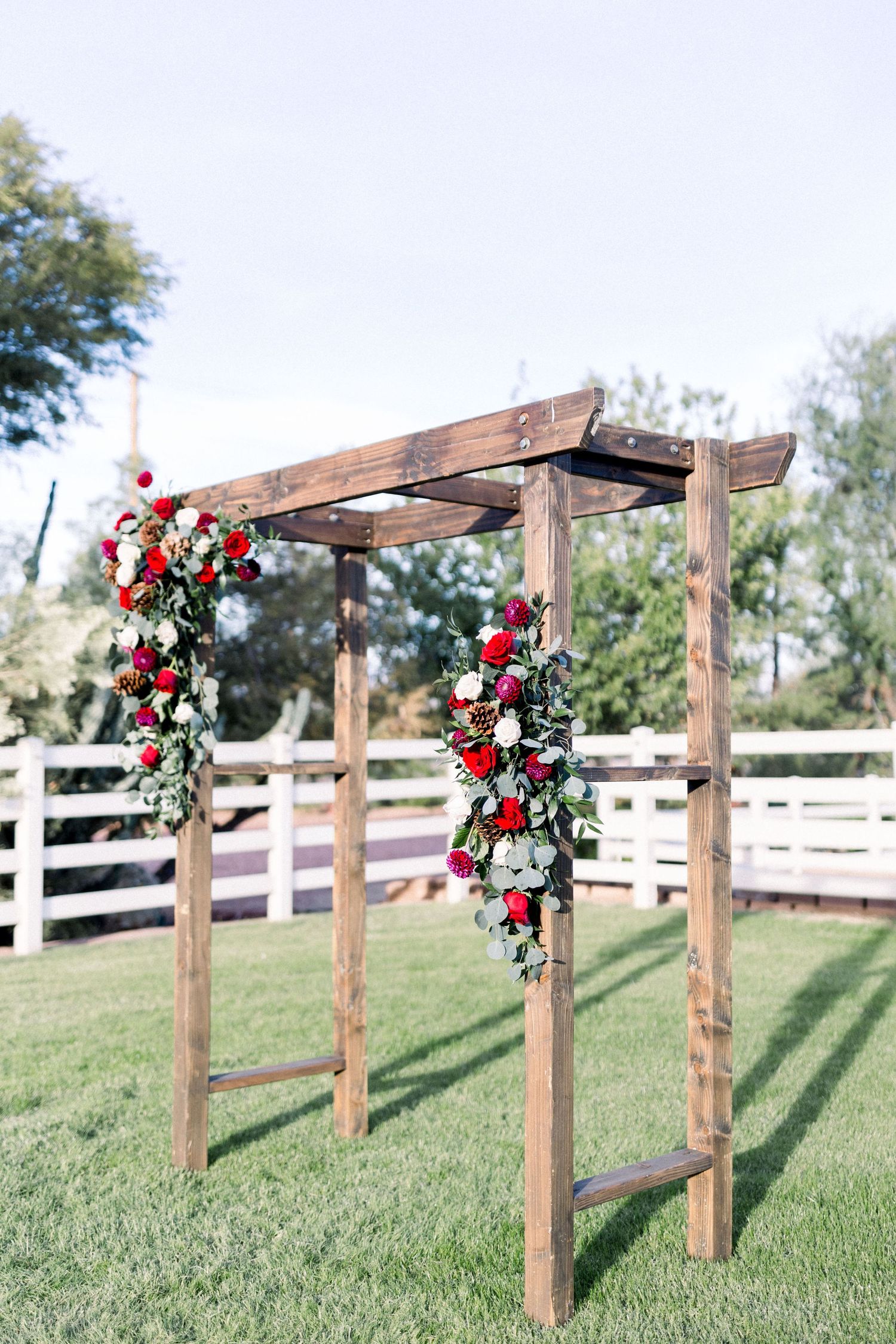 Rustic wooden wedding arch decorated with red and white flowers against a white fence backdrop.