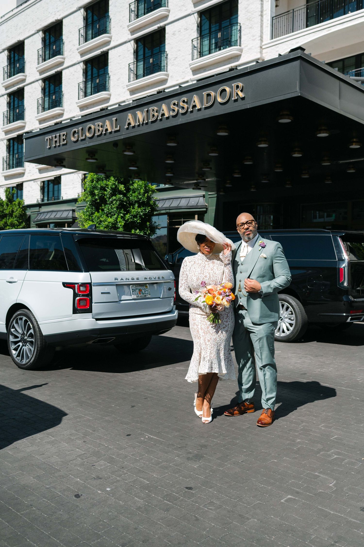 A couple in wedding attire poses outside The St. Regis Ambassador hotel entrance with luxury cars in view.