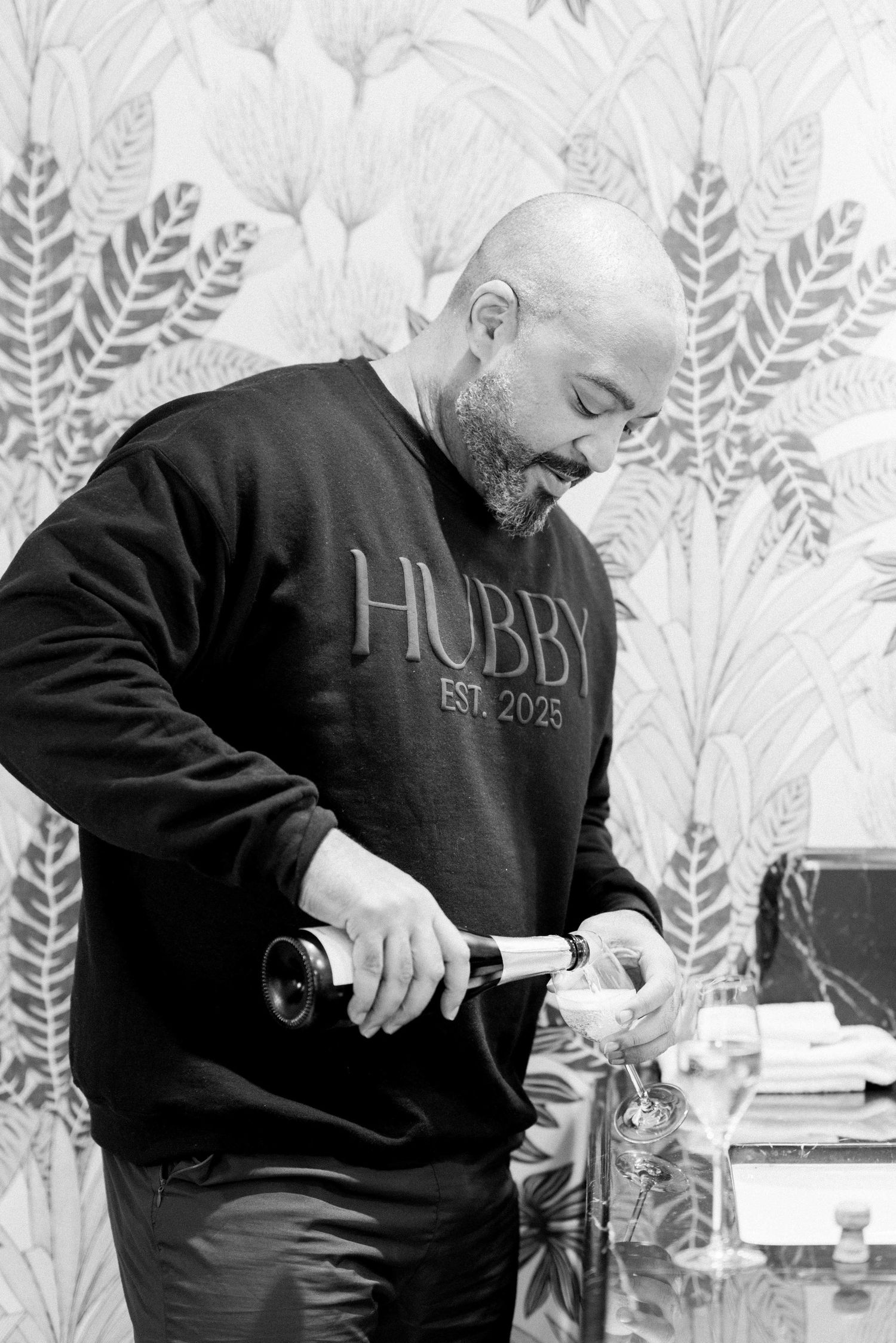 Person wearing a black 'HUBBY EST. 2023' sweatshirt pours a drink at an event with decorative wallpaper in background.
