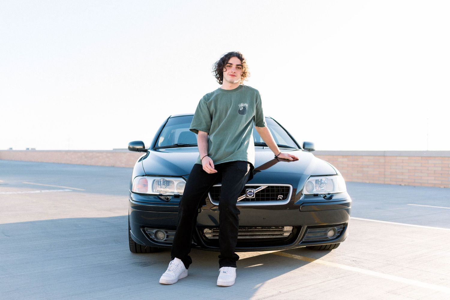 Person in casual attire leaning against black Volvo car on rooftop.