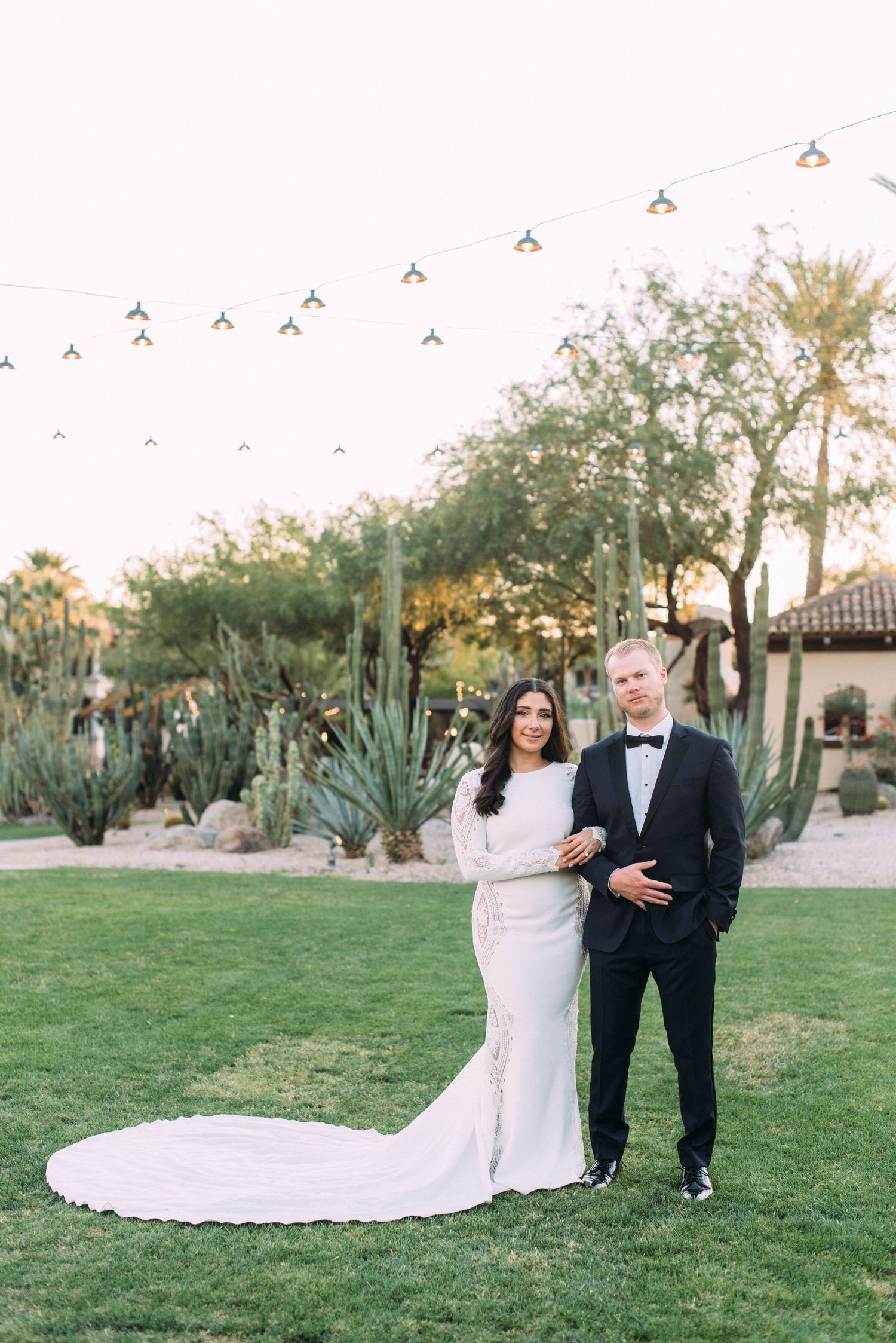 A wedding couple poses on manicured grounds with string lights and desert landscaping.
