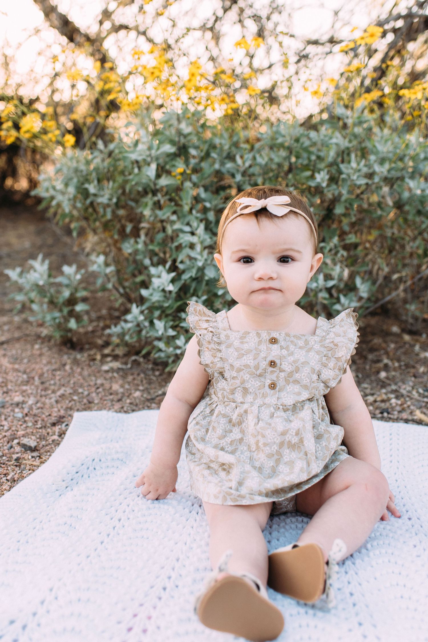 Baby wearing floral dress and bow sits on white blanket outdoors near flowering bushes.