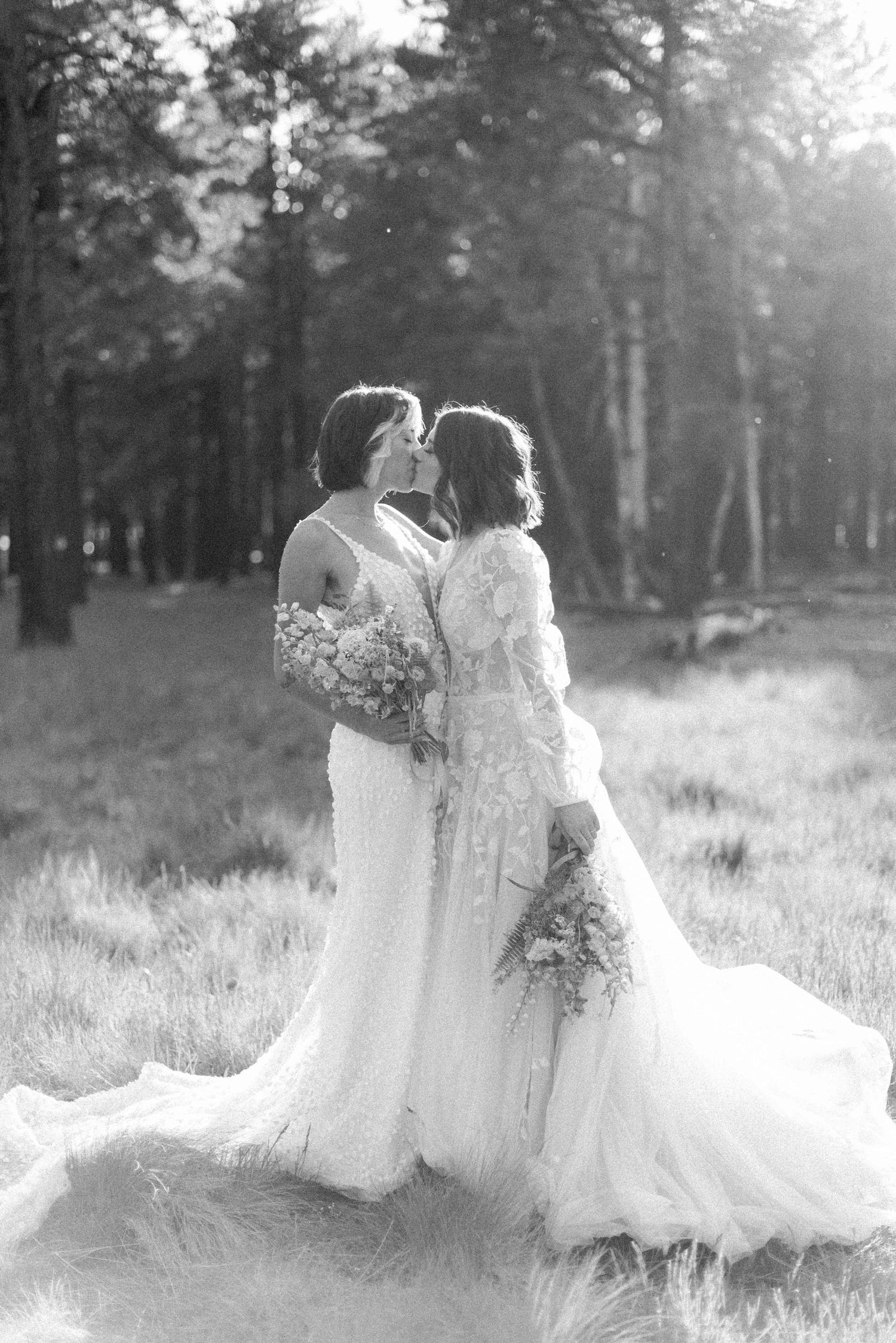 Black and white portrait of two brides in lace gowns embracing in a forest setting.