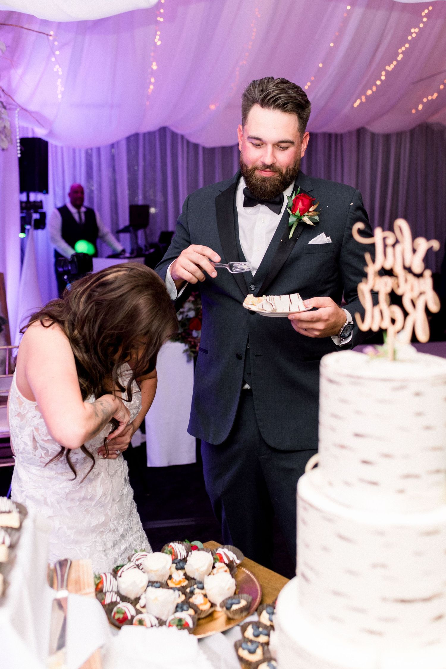 Newlyweds at their wedding reception sharing a playful moment while cutting their wedding cake in a purple-lit tent venue.