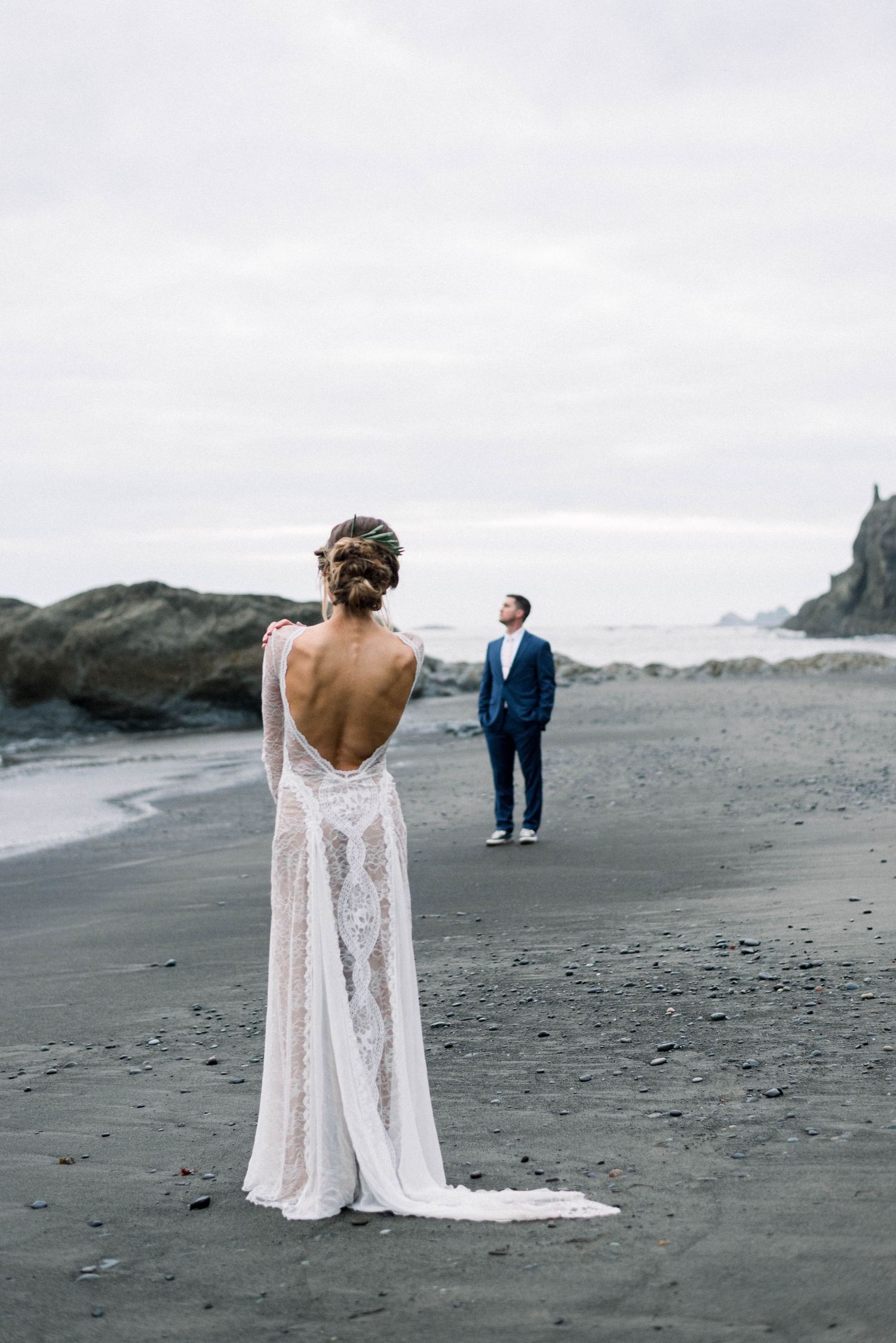A bride in a backless lace wedding dress stands on a moody black sand beach with cliffs in the distance.