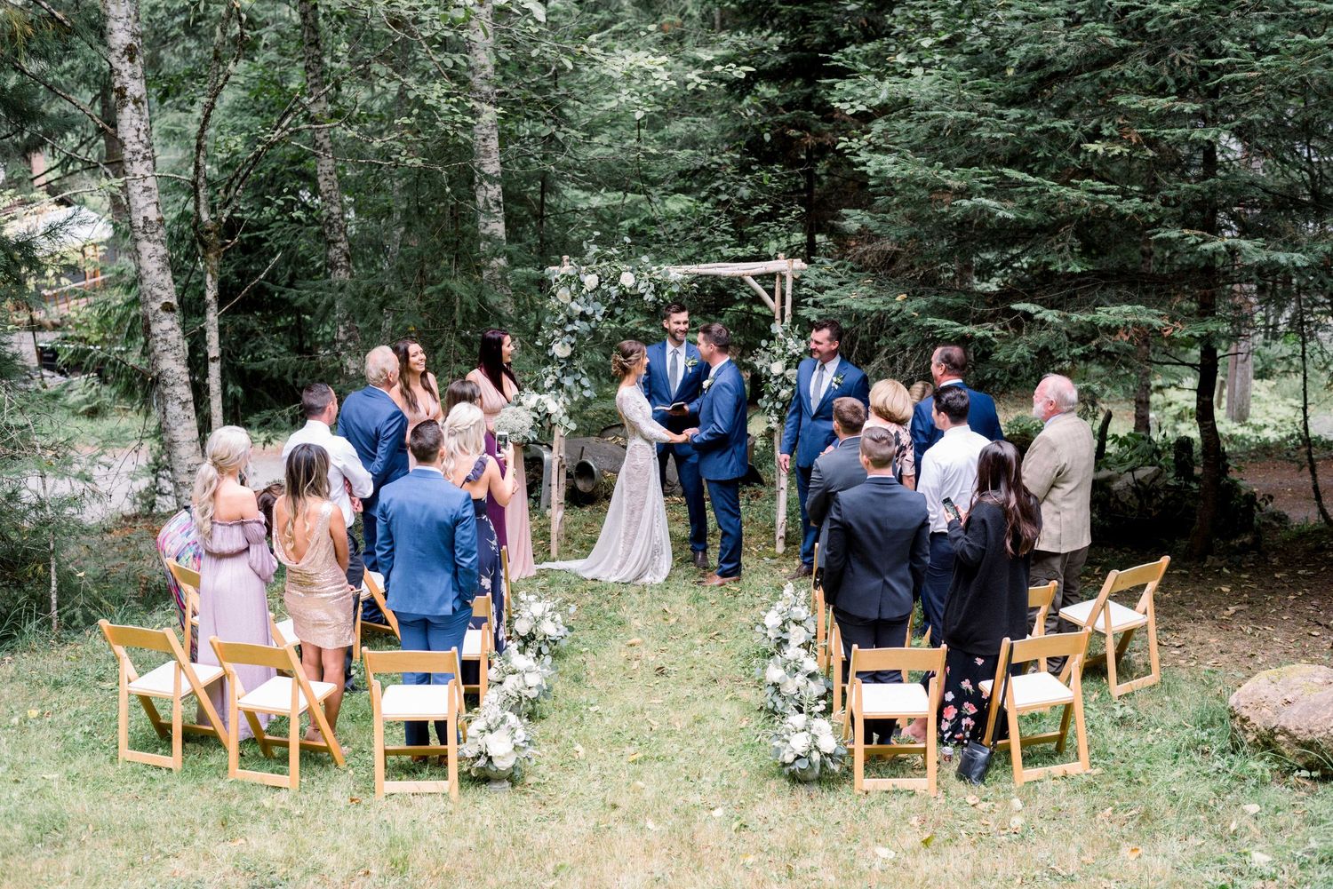 An intimate forest wedding ceremony with guests seated on wooden chairs among pine trees.