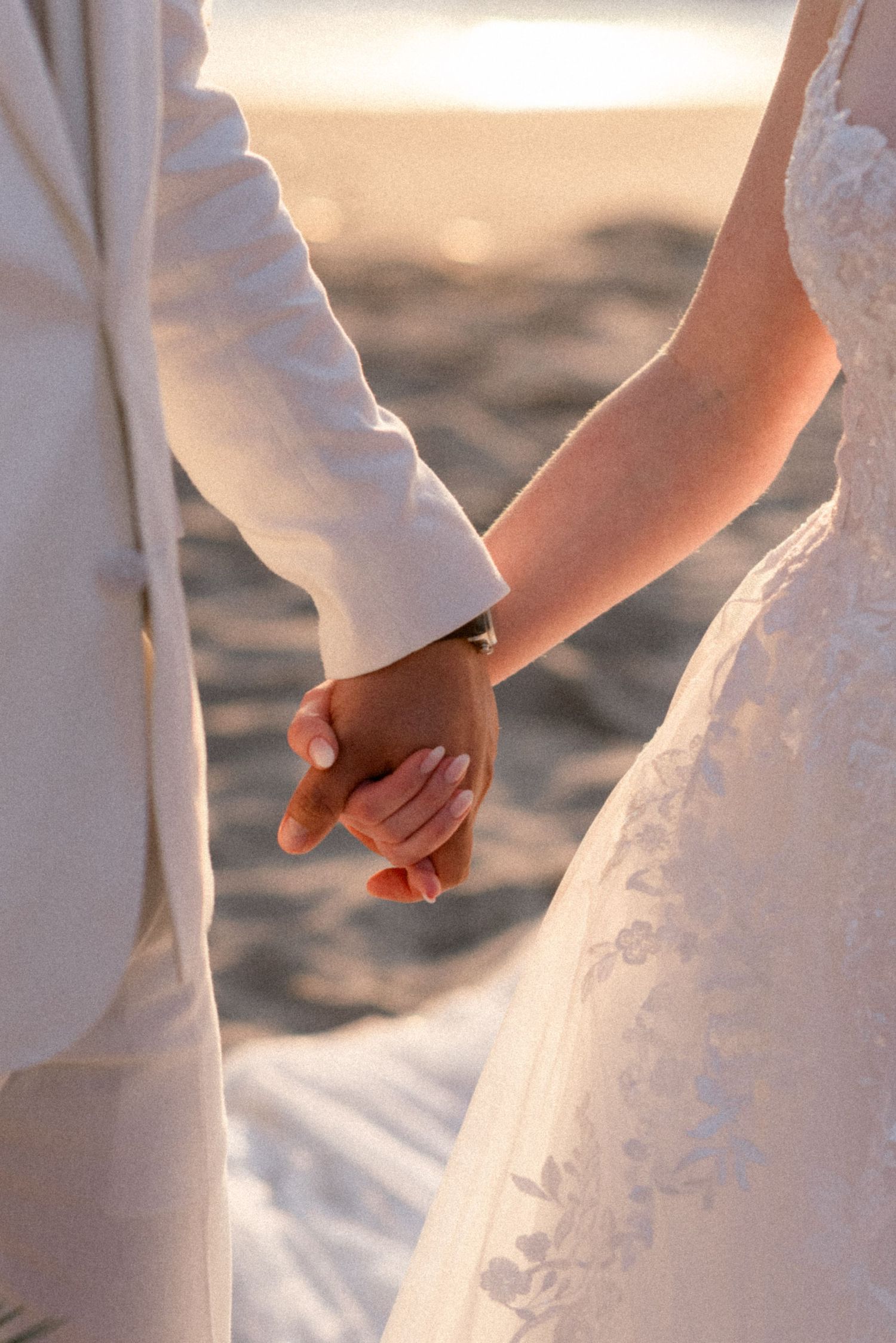 Close-up of newlyweds holding hands on beach with white dress and suit details.