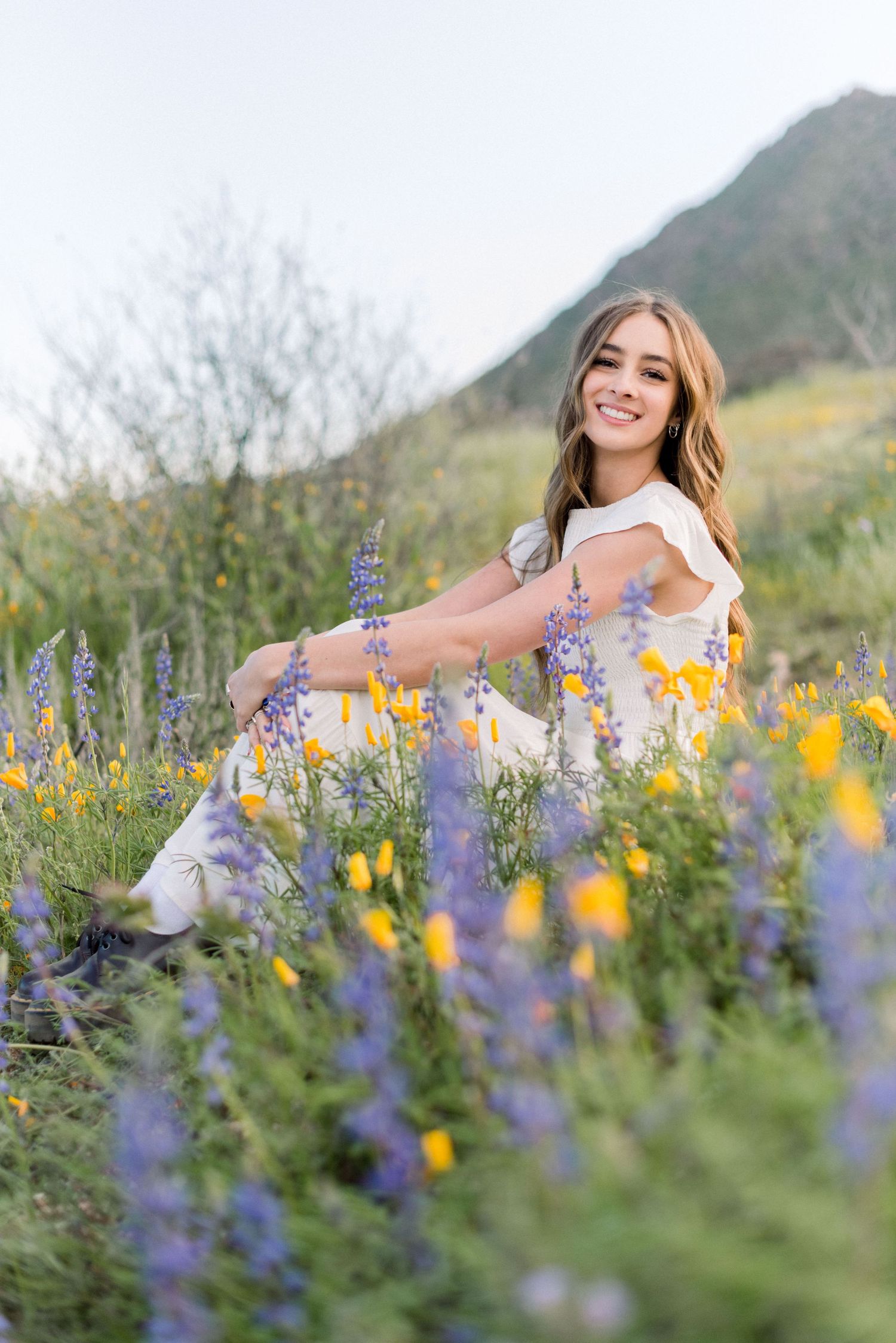 A woman sits among vibrant purple and yellow wildflowers against a mountain backdrop.