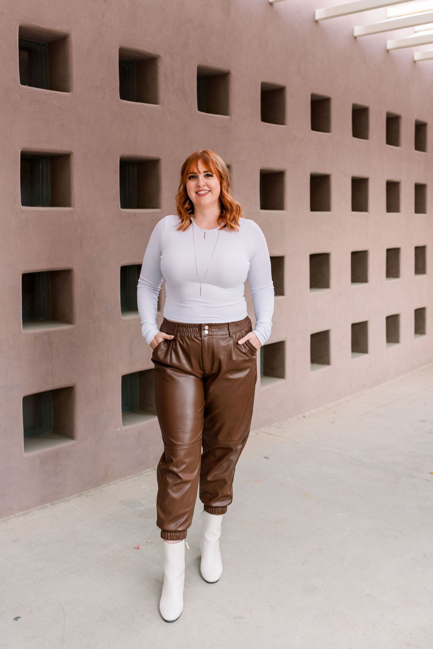 Fashion model wearing white top and brown leather pants poses against geometric wall with square cutouts.