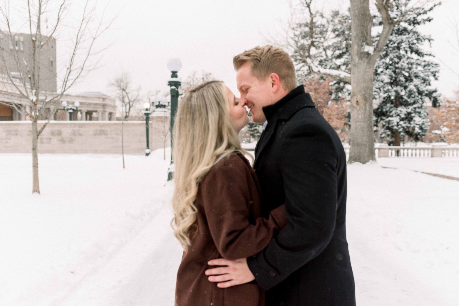 Couple sharing intimate winter kiss with snow-covered trees in background.