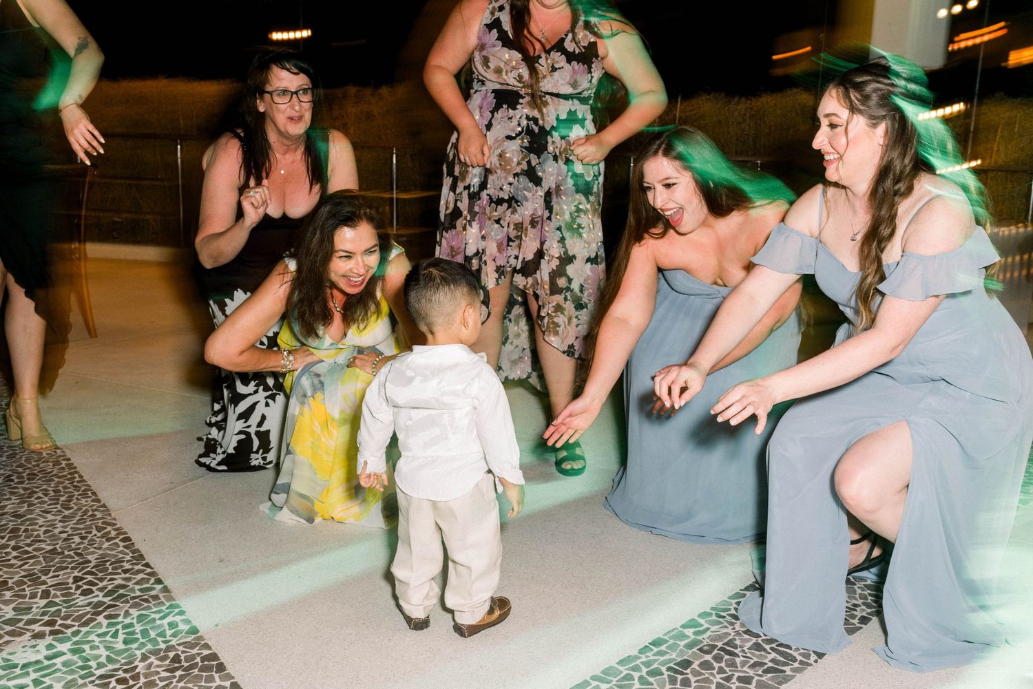 A little boy in white surrounded by guests in formal dresses dancing at an evening celebration.