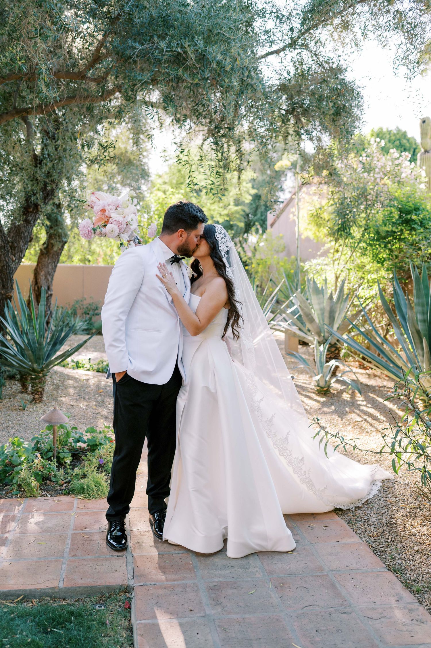 A bride and groom share a romantic kiss in a lush desert garden, with agave plants and olive trees surrounding them.