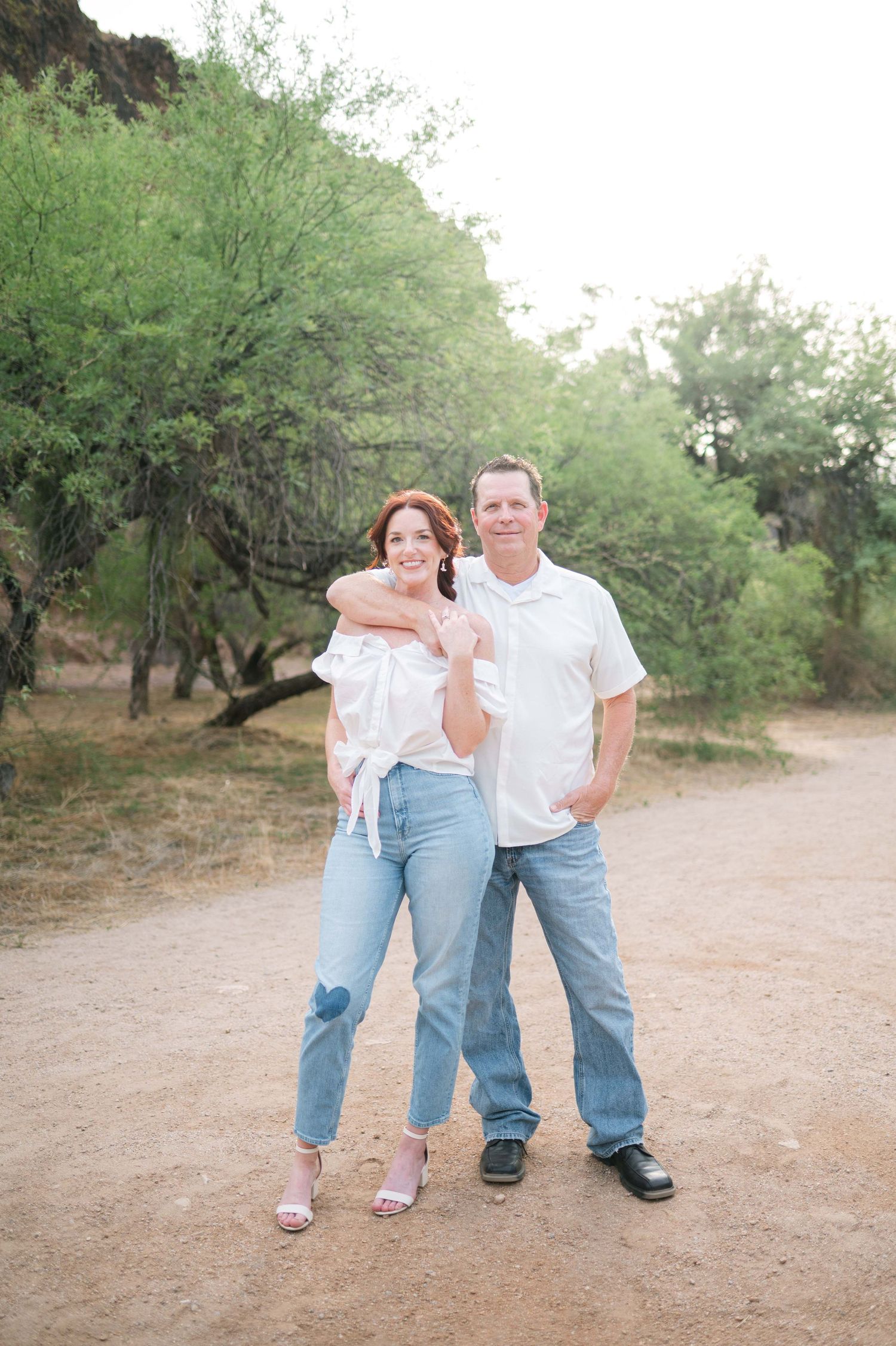 A couple in casual white shirts and jeans poses together on a desert path surrounded by green desert trees.
