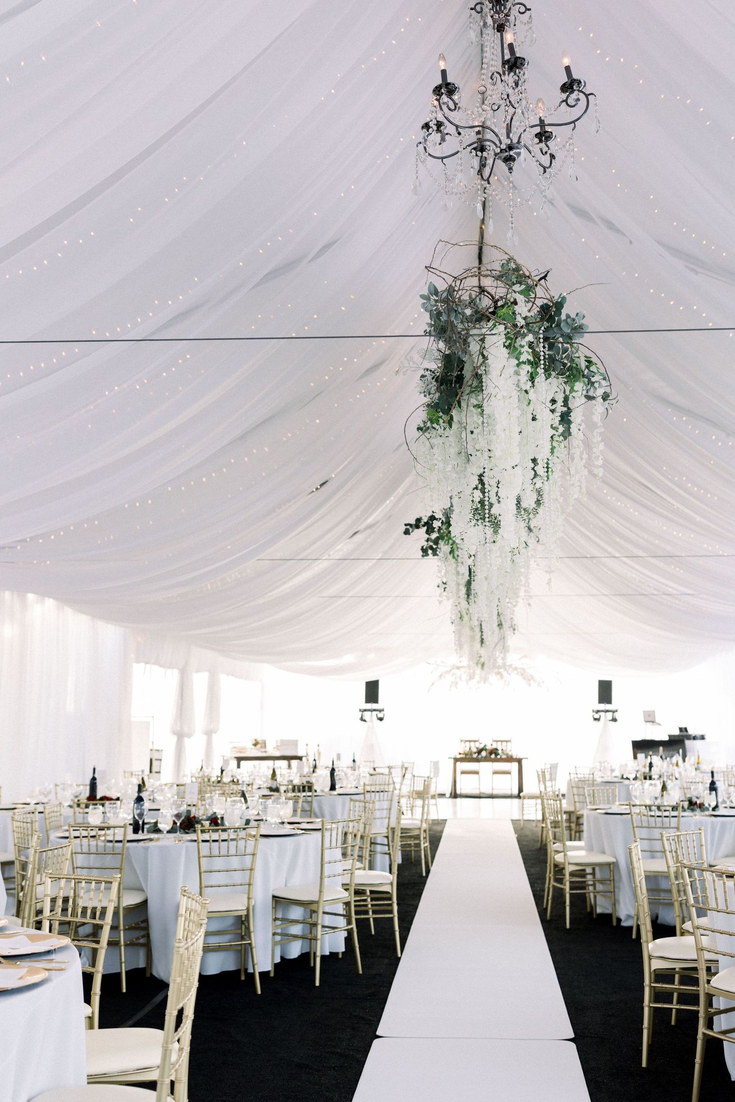 Elegant white tent reception with chandelier and floral arrangements hanging from draped ceiling.